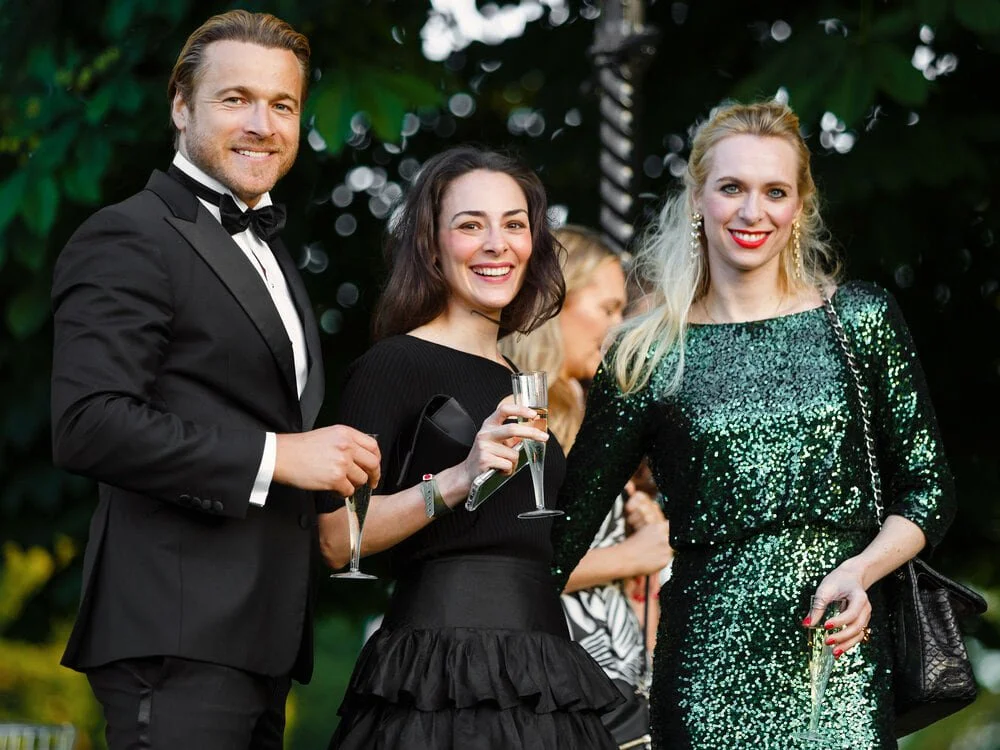 Three guests dressed in formal evening wear holding drinks and posing together during a corporate networking event.
