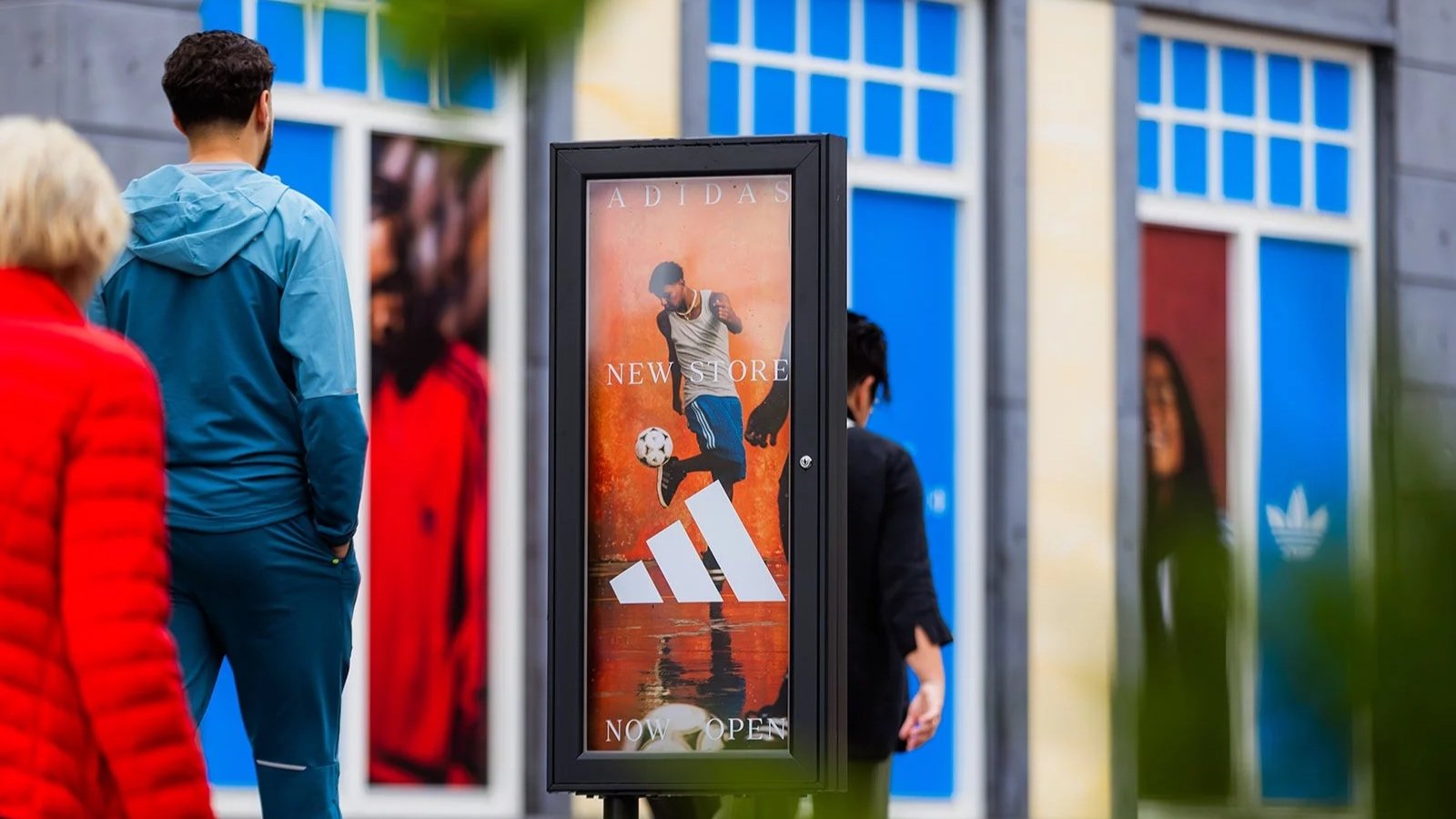 Adidas new store opening sign outside the Roermond shop, with passersby and blue branded window displays behind it.