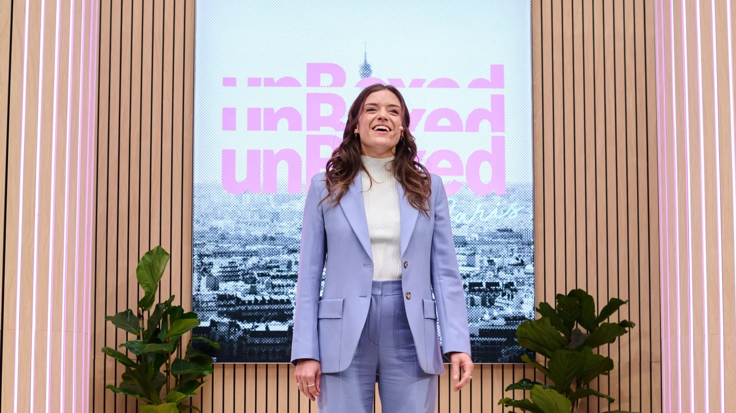 Smiling speaker in a lilac suit wearing a headset microphone in front of an Amazon Unboxed Paris backdrop.
