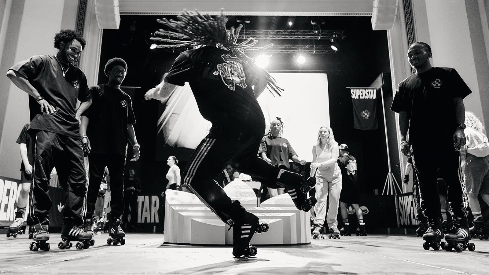 Black-and-white photo of a roller skater performing with one leg raised as others watch at the Adidas x Offspring Superstar Rollerskates event.