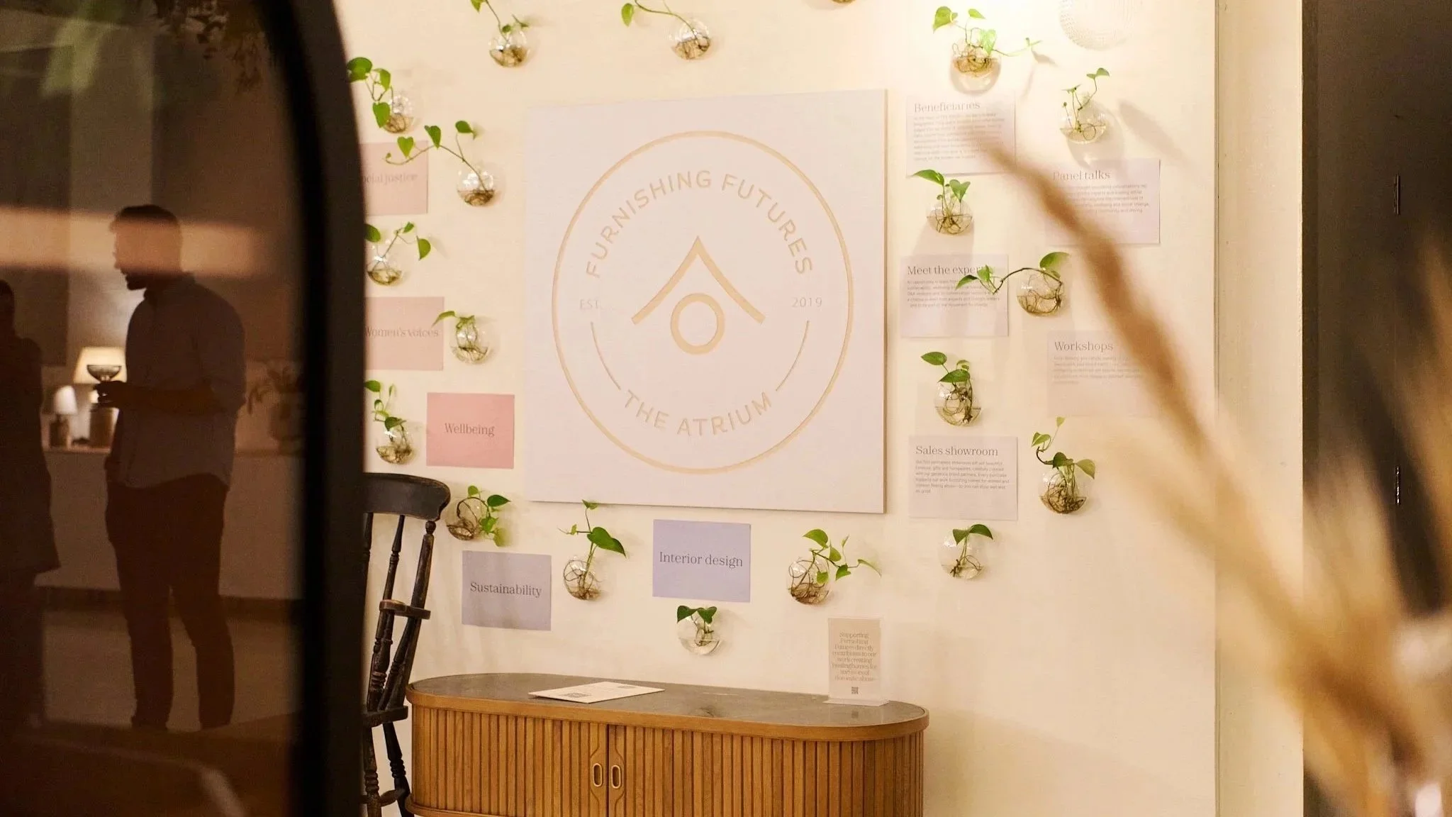 Furnishing Futures logo surrounded by small wall-mounted plants at The Atrium in East London, with a guest holding a drink reflected in glass.