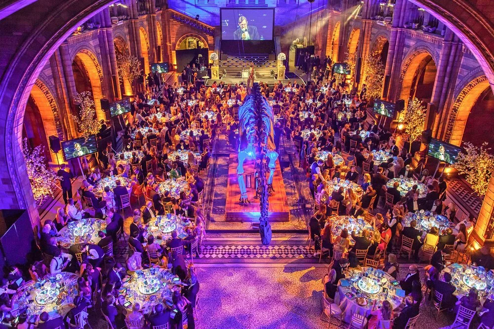 Guests seated at tables during a large awards dinner inside a historic museum venue with dramatic lighting and a dinosaur skeleton in the centre of the room.