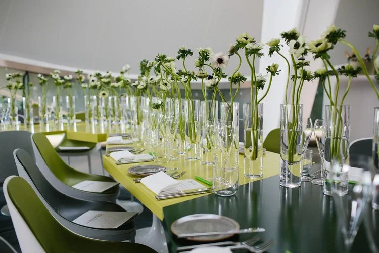 Long table set for a corporate event with green chairs, glass vases of white flowers, place settings, and natural light.