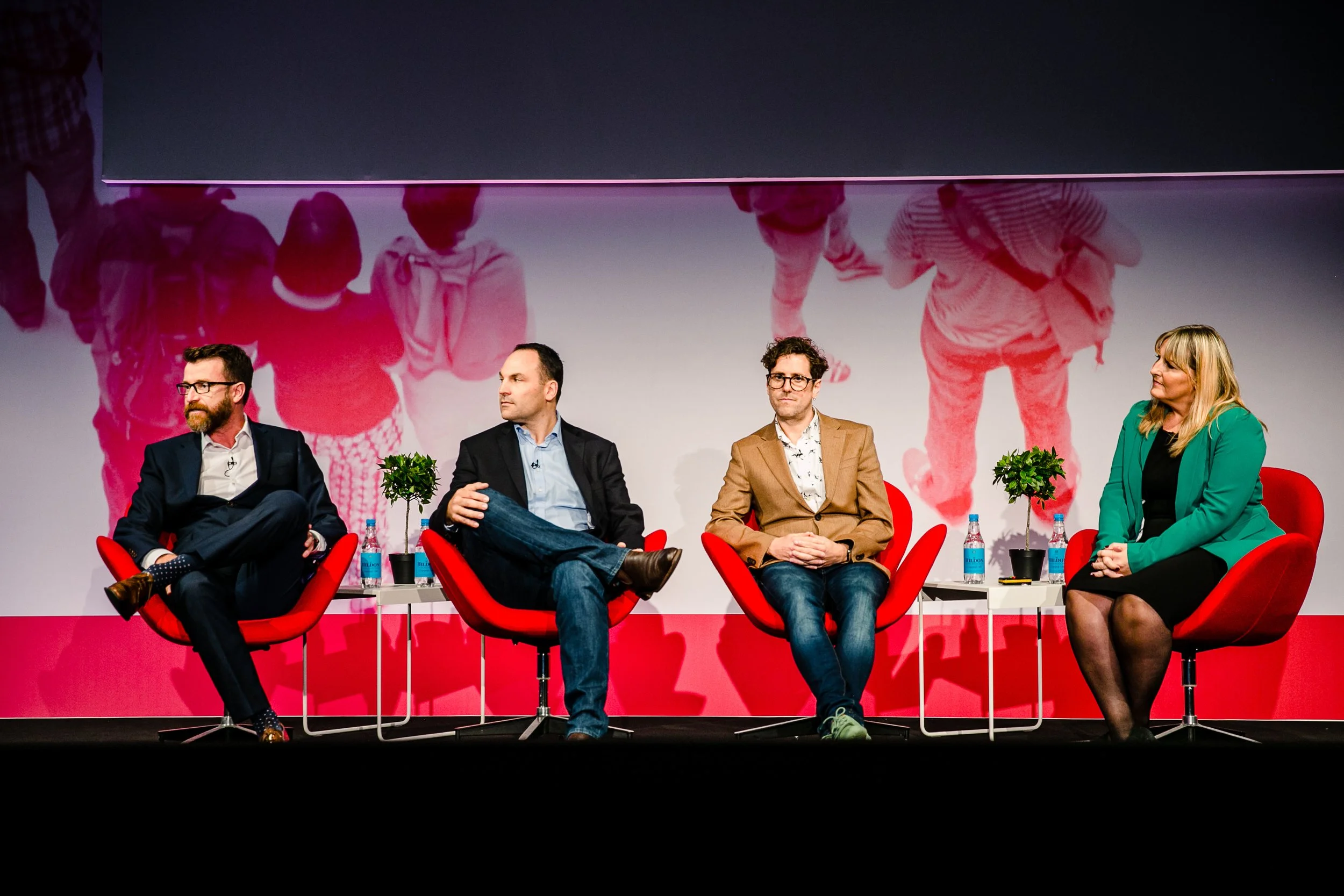 Four speakers seated on red chairs on a stage during a panel discussion, with a large screen backdrop behind them.