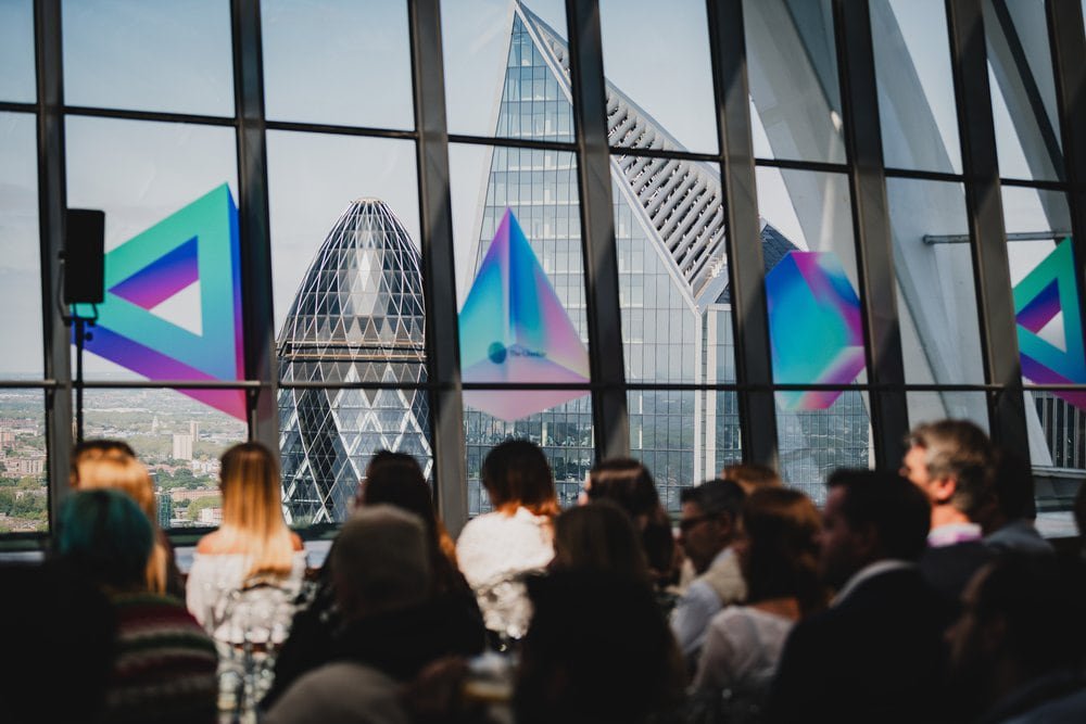 Audience seated at a conference event inside a glass venue overlooking the Gherkin and City of London skyline.