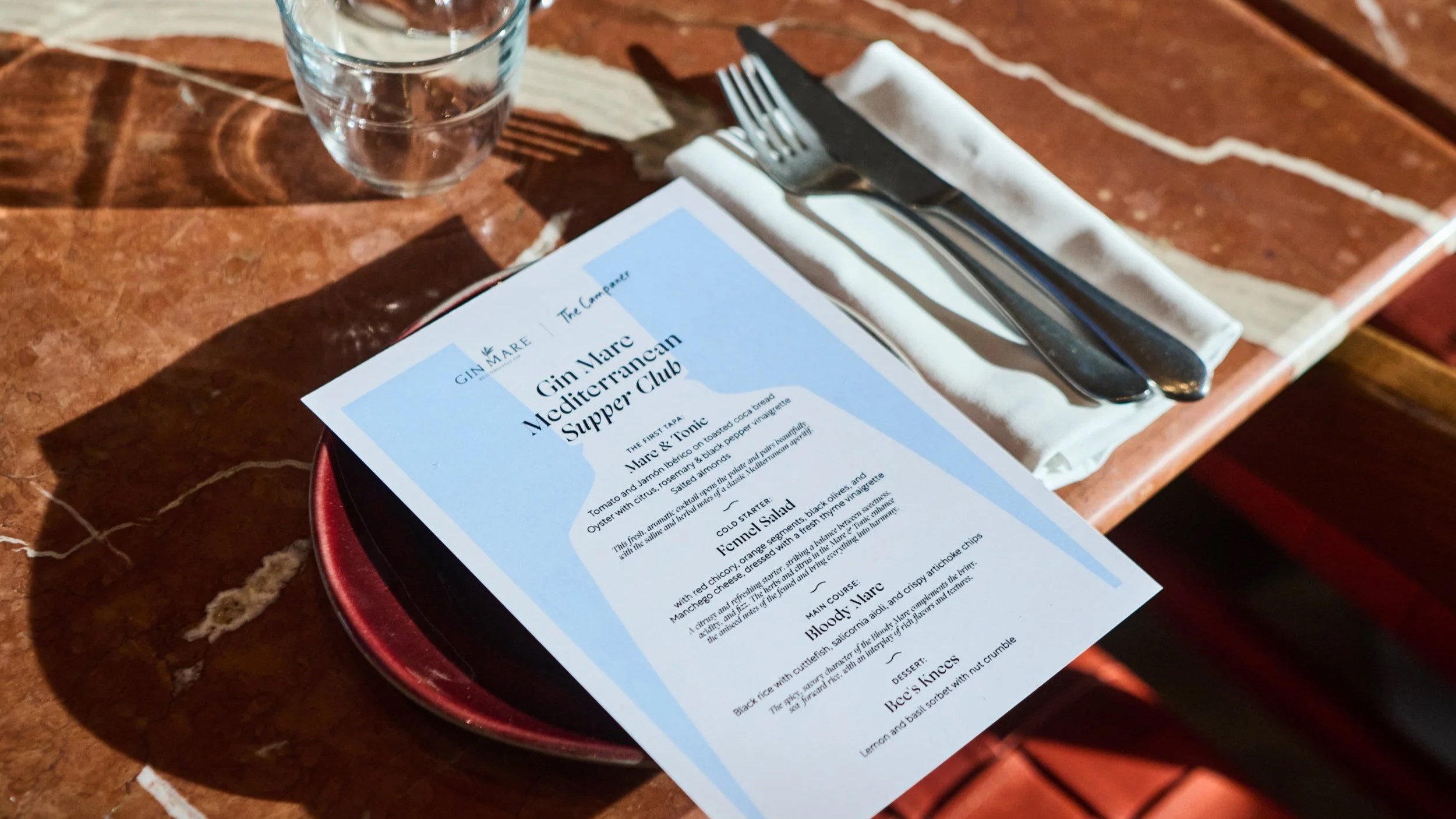Gin Mare Mediterranean Supper Club menu on a red plate beside cutlery and a water glass at The Campaner, London.