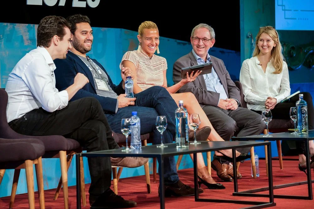 Panel discussion taking place on stage with five speakers seated in front of a conference audience.