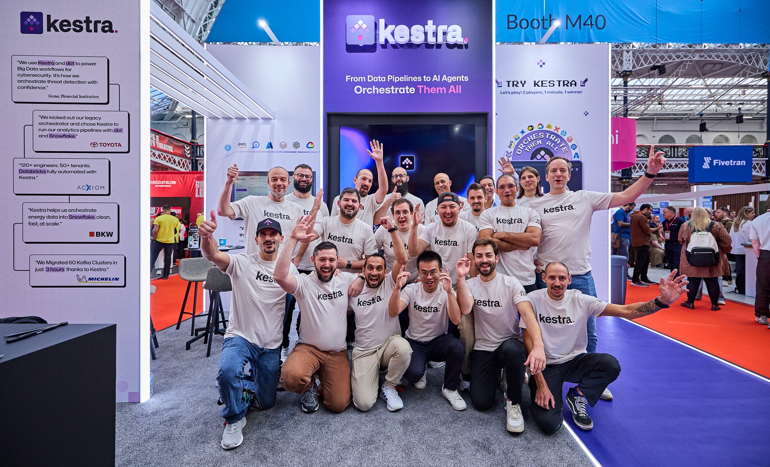 Group of people wearing matching white T-shirts standing and kneeling together at an exhibition trade show stand with signage behind them.