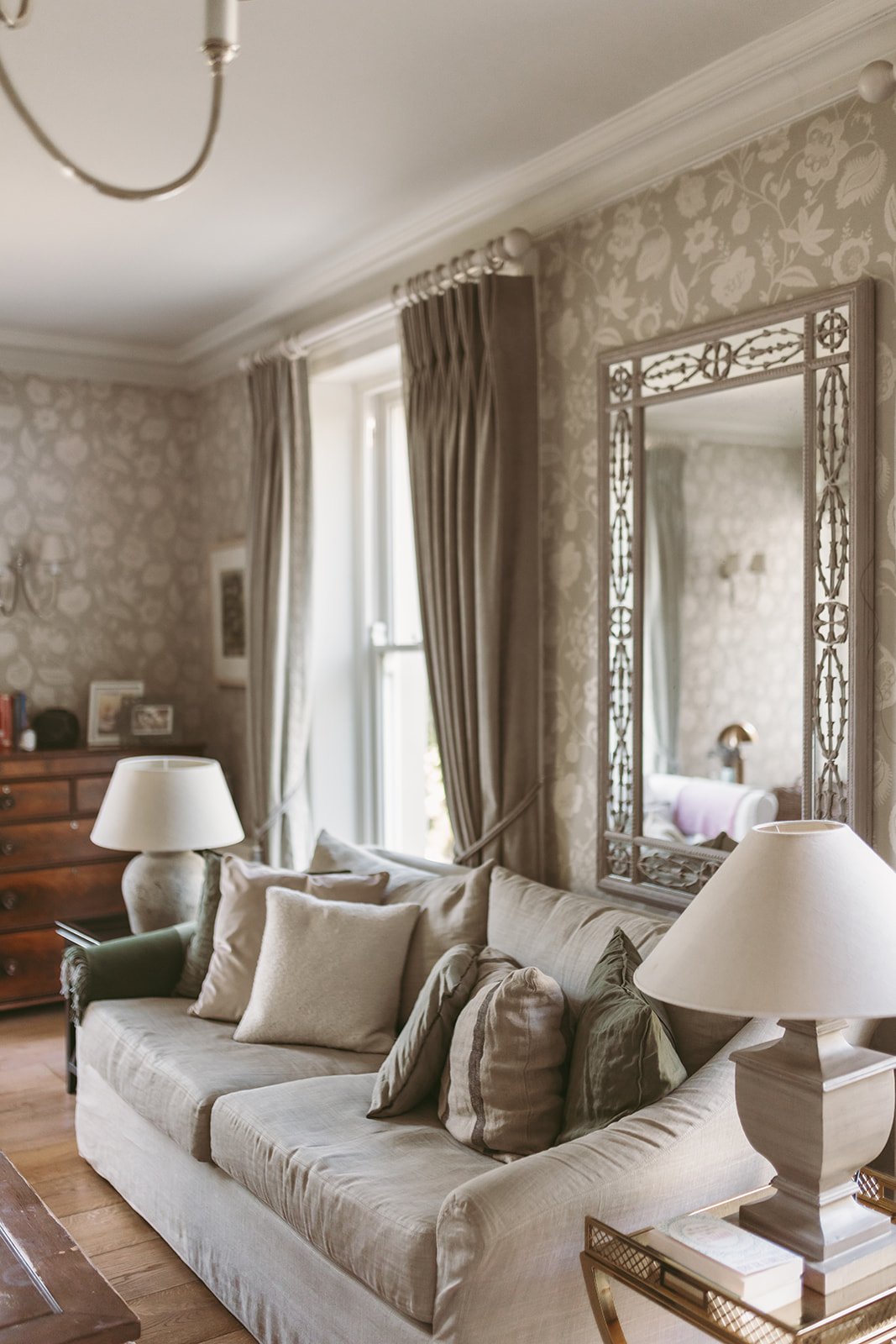 A cozy living room with beige patterned wallpaper, a cream sofa with multiple pillows, two lamps, a wooden dresser, and a large decorative mirror on the wall.