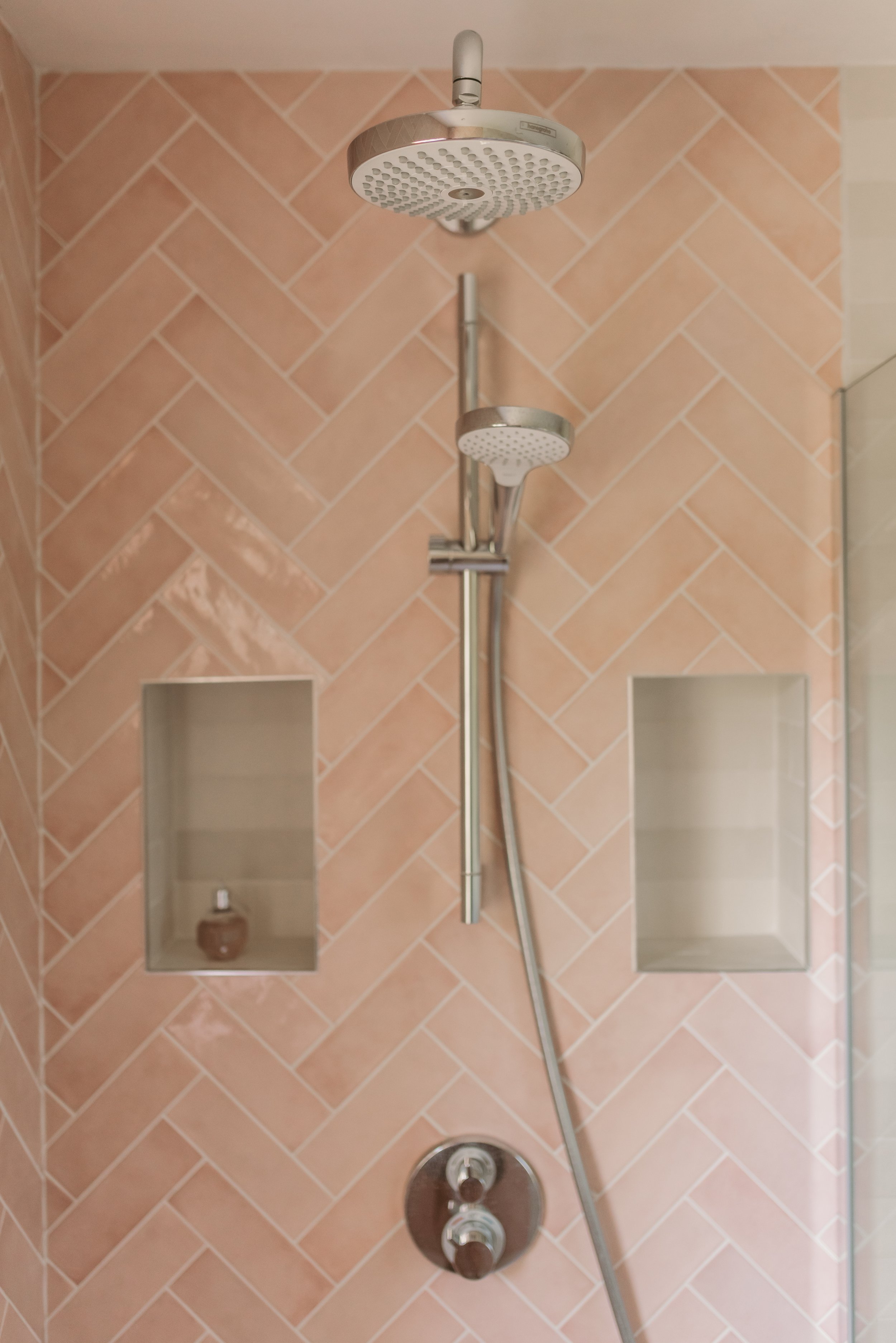 Shower with pink herringbone tile wall, two showerheads, and two recessed shelves with a bottle in one of them.