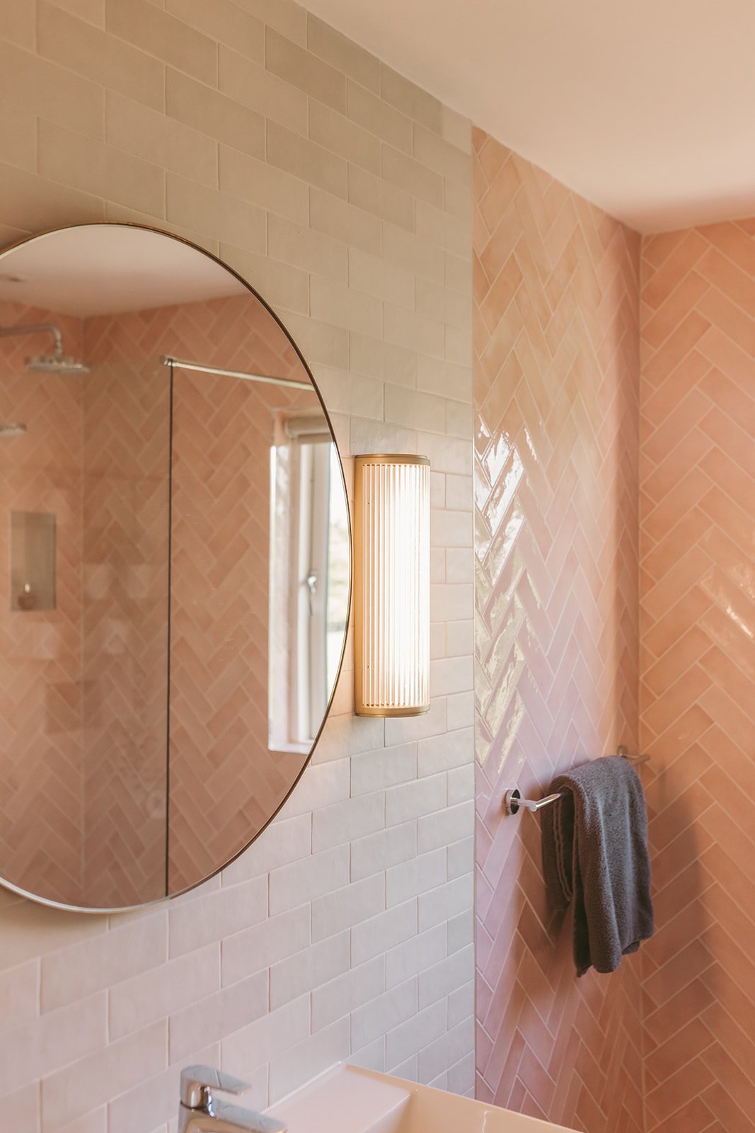 A bathroom with a round mirror, wall-mounted light fixture, pink tiled walls, a small window, and a gray towel hanging on a towel rod.