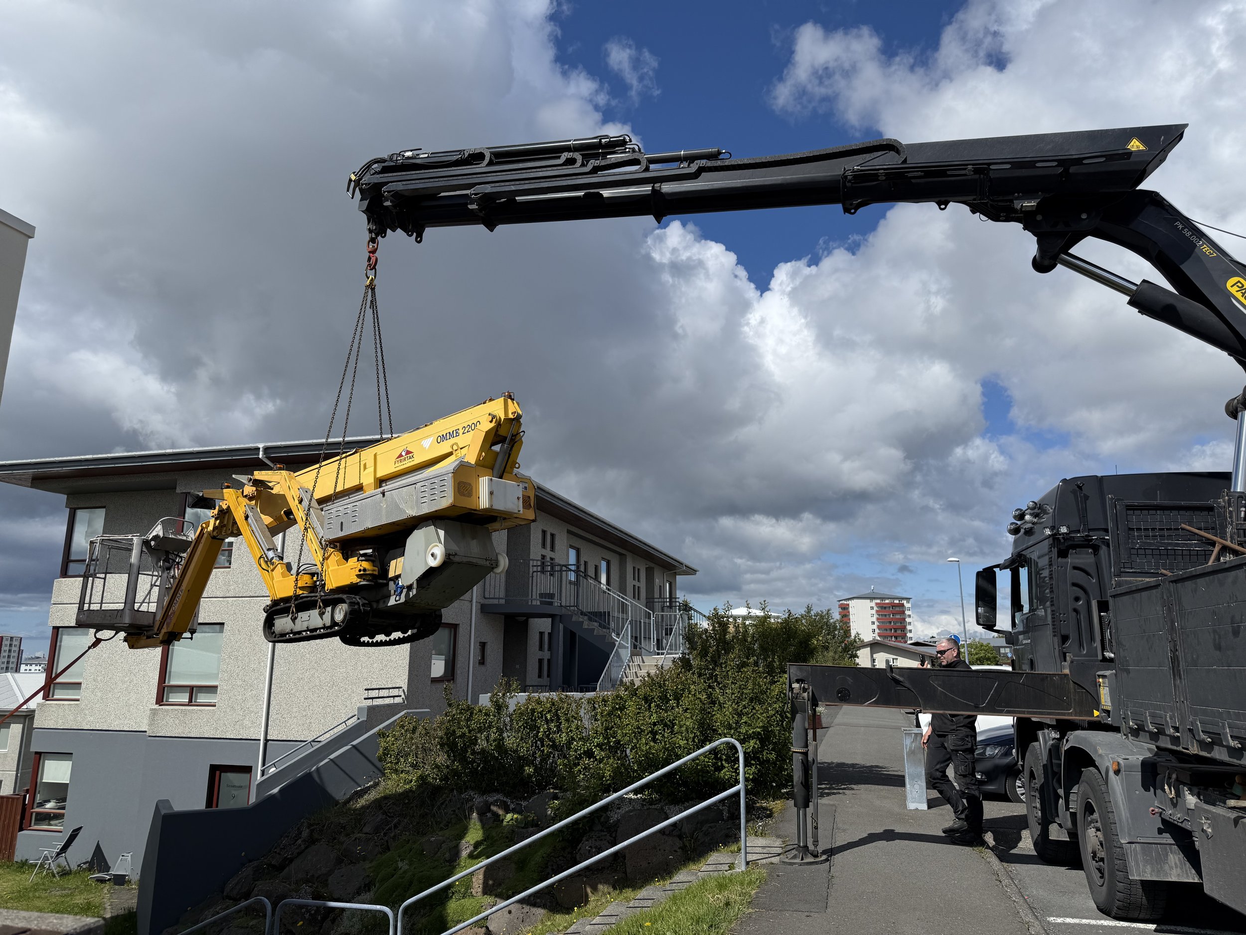 Construction workers using a crane and a lift to install or repair a utility cable on a building exterior during daytime with a partly cloudy sky.