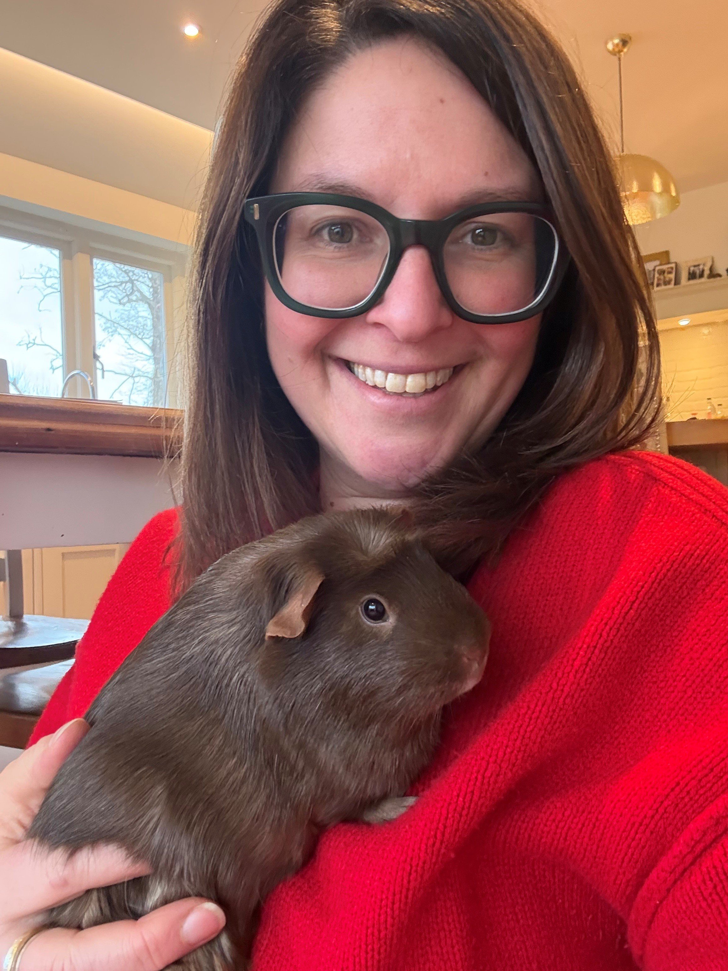 A smiling white woman with brown hair wearing green glasses & a red jumper holding a dark brown guinea pig