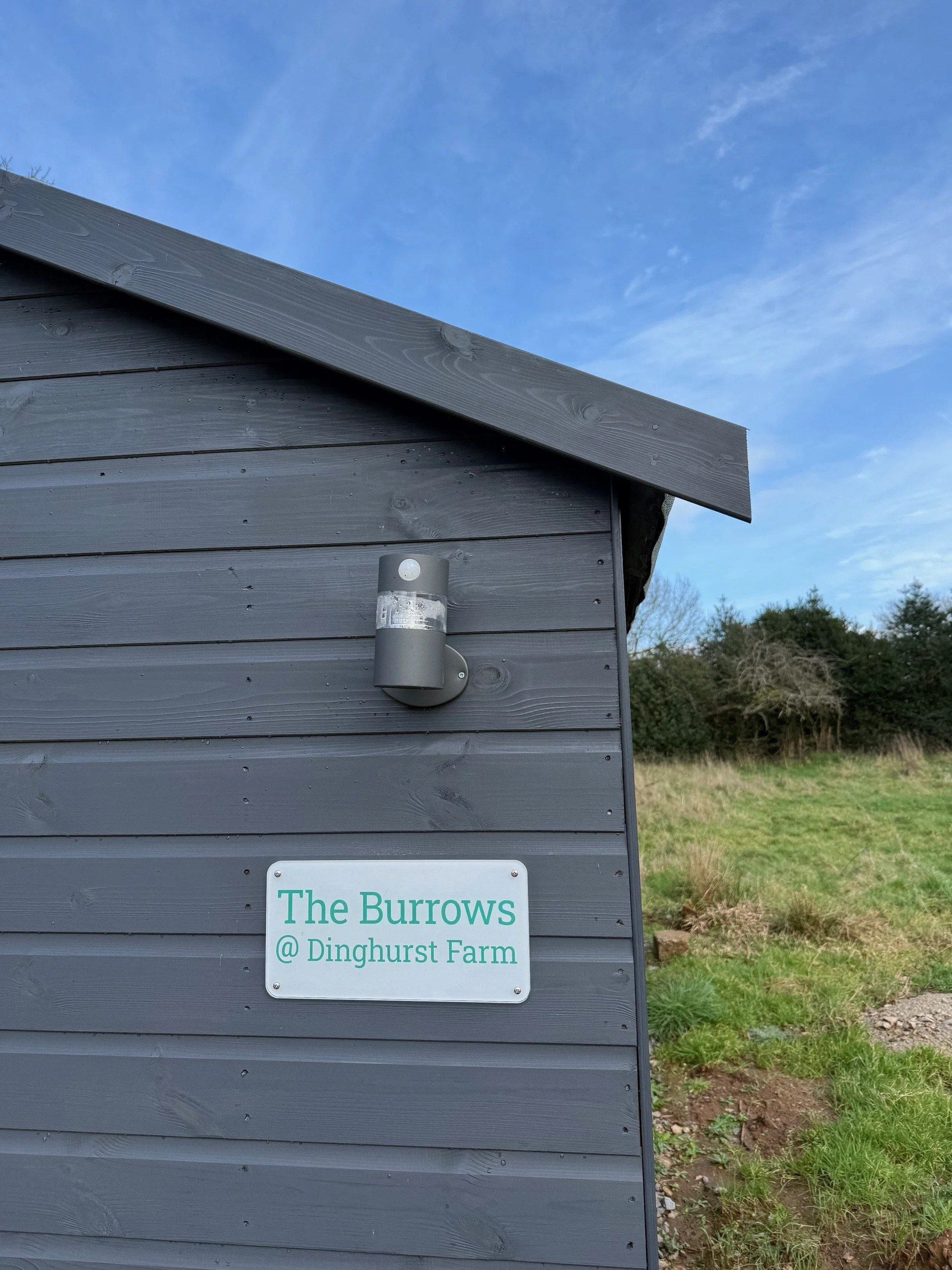 A grey painted shed with a white sign with green text that reads The Burrows @ Dinghurst Farm