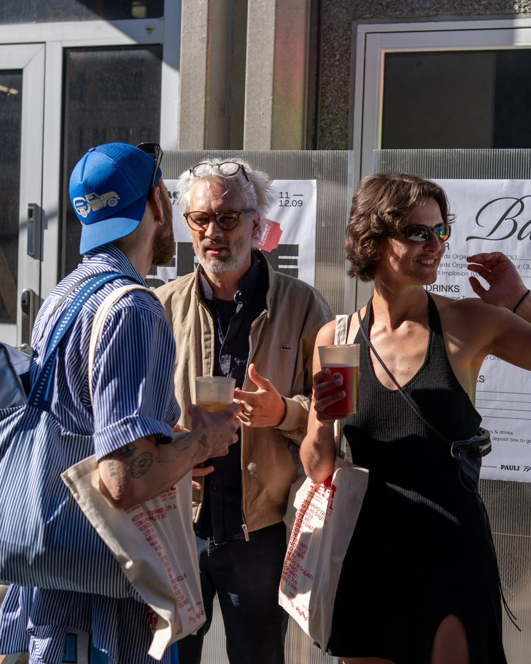 Three people are standing outdoors, engaged in conversation, with drinks in hand. They are dressed casually, and the scene is sunlit with a sign behind them.