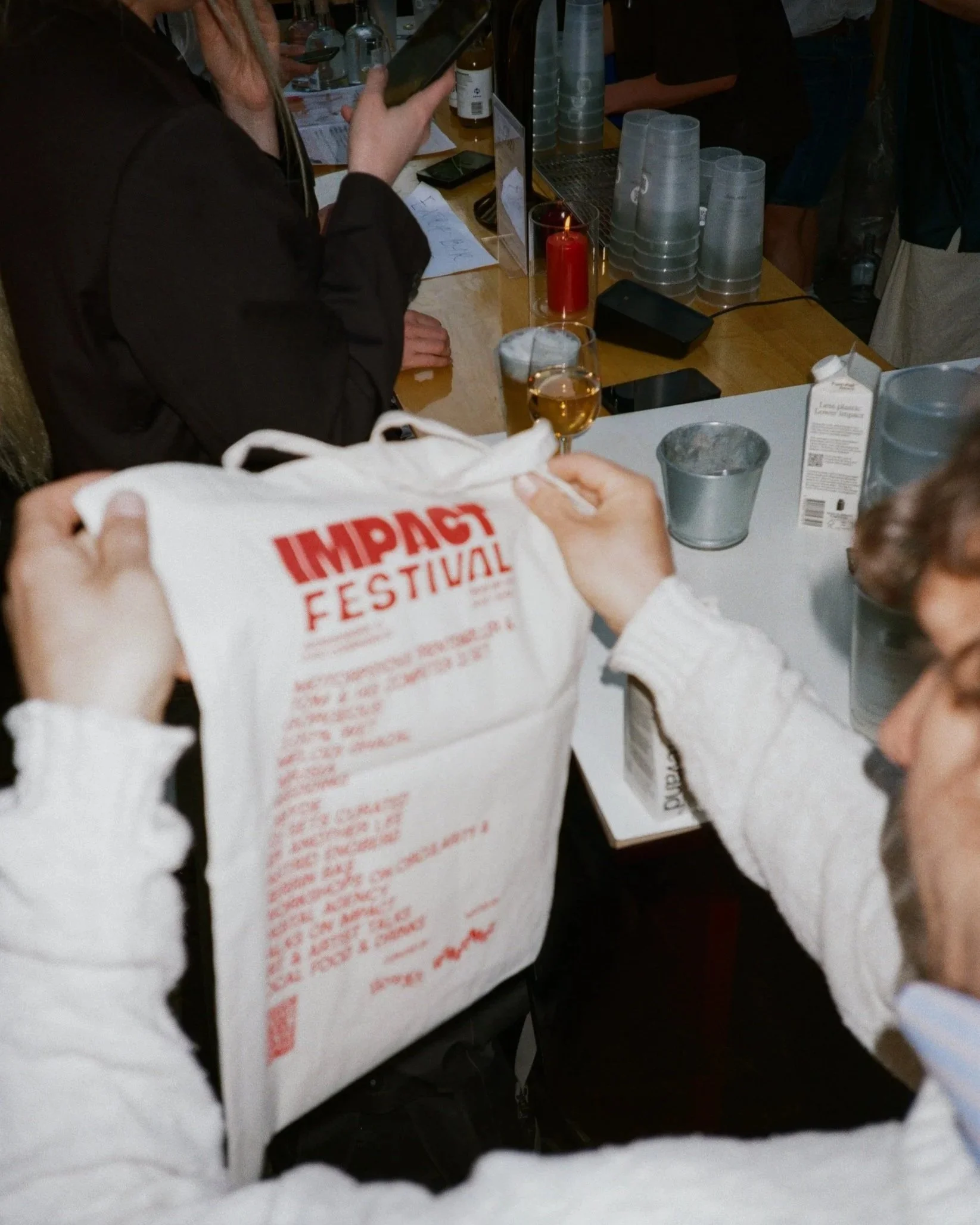 A person holding a white bag with red text that reads 'IMPACT FESTIVAL' in a crowded bar or restaurant setting, with what appears to be a glass of beer or cider on the counter and various bottles and cups in the background.