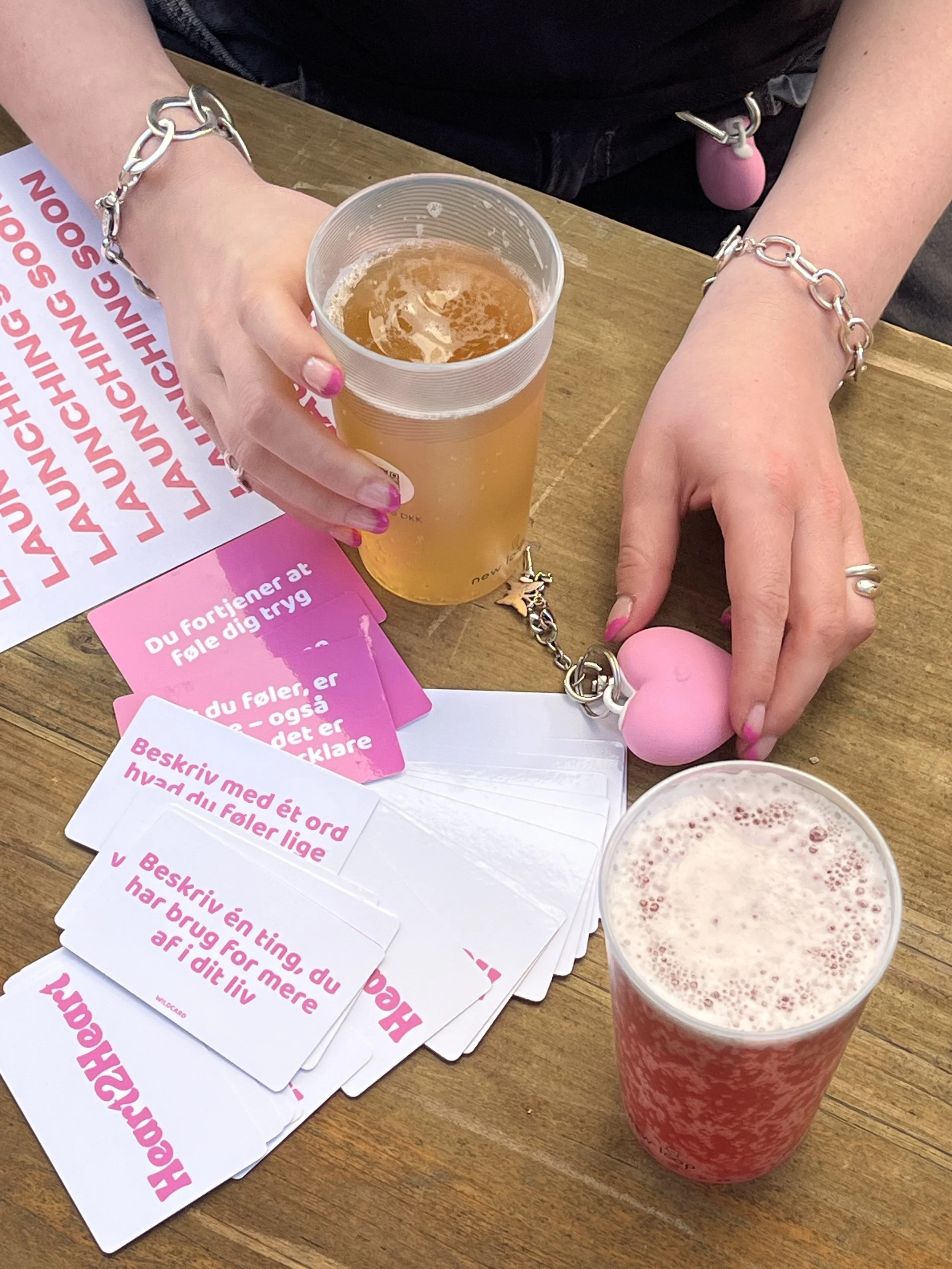 Two drinks on a wooden table, one appears to be beer in a clear plastic cup, the other is pink and appears to be a berry smoothie or juice. There are also several pink and white papers with text in Danish, along with a chain bracelet and a pink heart-shaped keychain on the table.