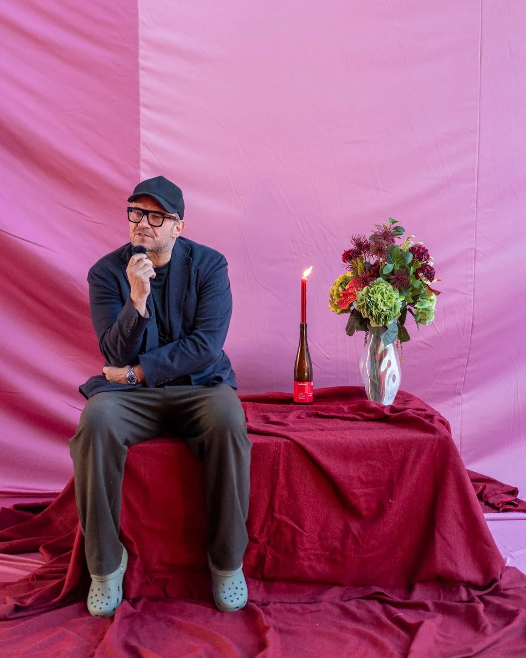 Thomas Asbæk sitting on a red draped table holding a microphone with a pink backdrop. Next to him are a tall red candle in a bottle-shaped holder and a white vase filled with a colorful arrangement of flowers.