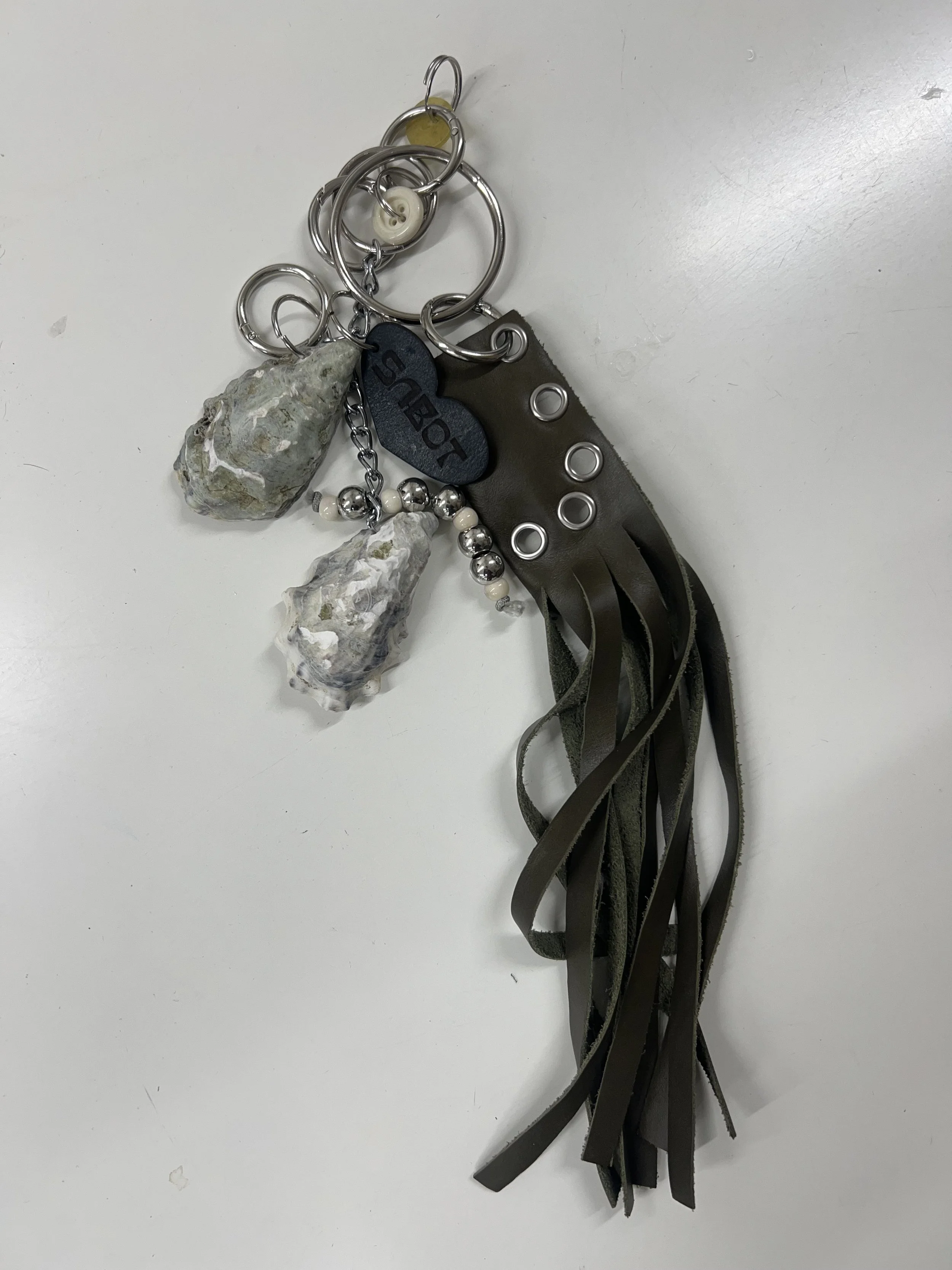 Keychain with oyster shells, a black heart-shaped tag reading 'SABOT' a metal bead chain, a leather strap, and metal rings on a white surface.