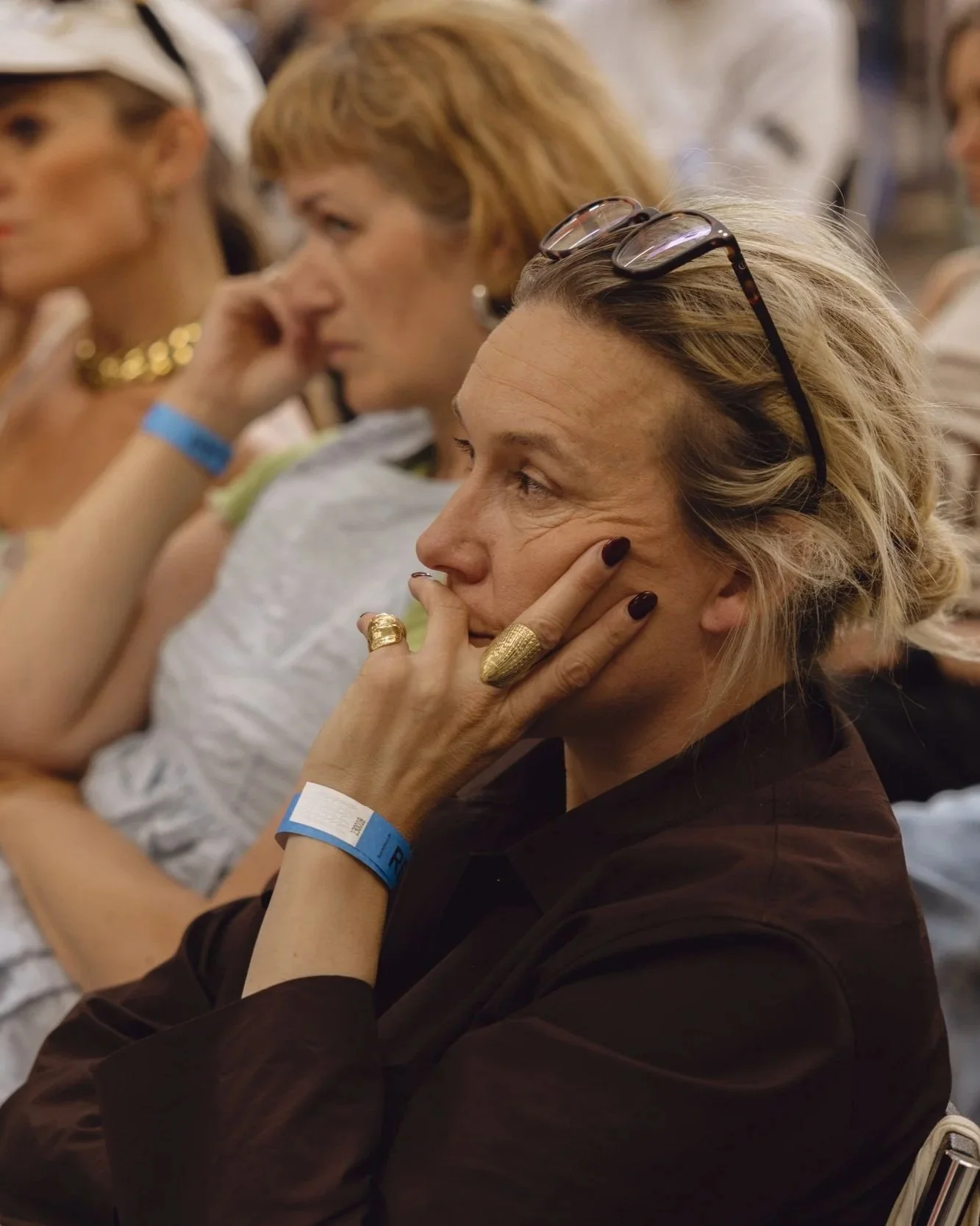 A woman with blonde hair and glasses resting on her head is sitting with her hand on her face, wearing a dark top and gold jewelry, at a crowded event.