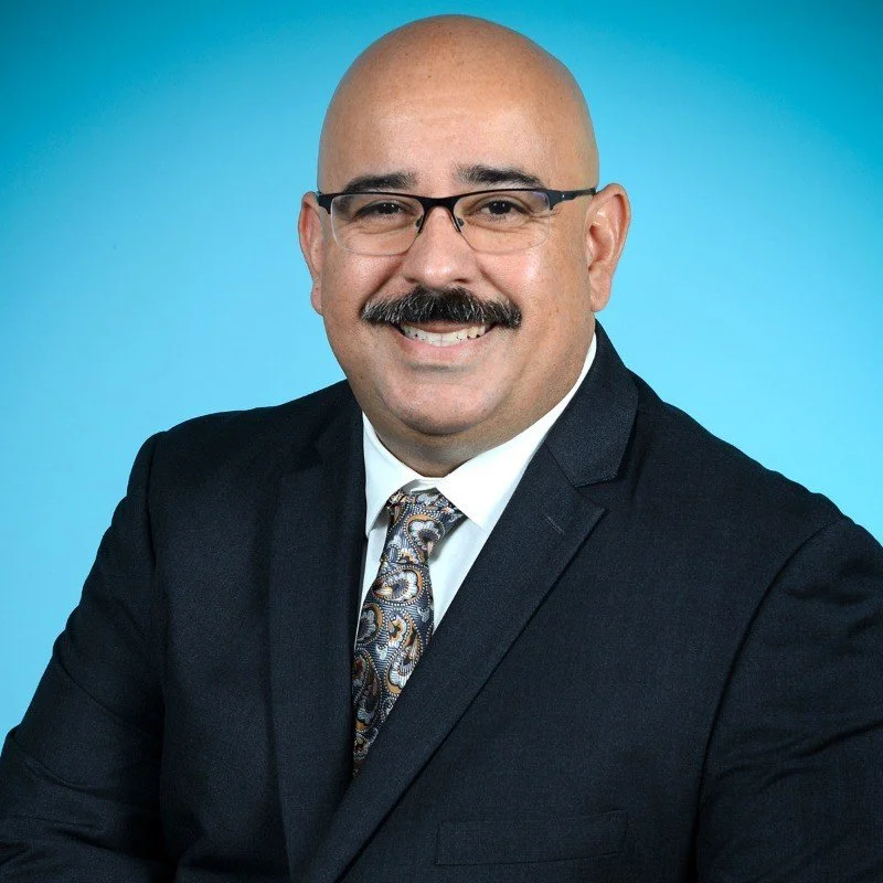 A professional man with glasses, a mustache, and a bald head, wearing a dark suit, white shirt, and patterned tie, smiling against a blue background.
