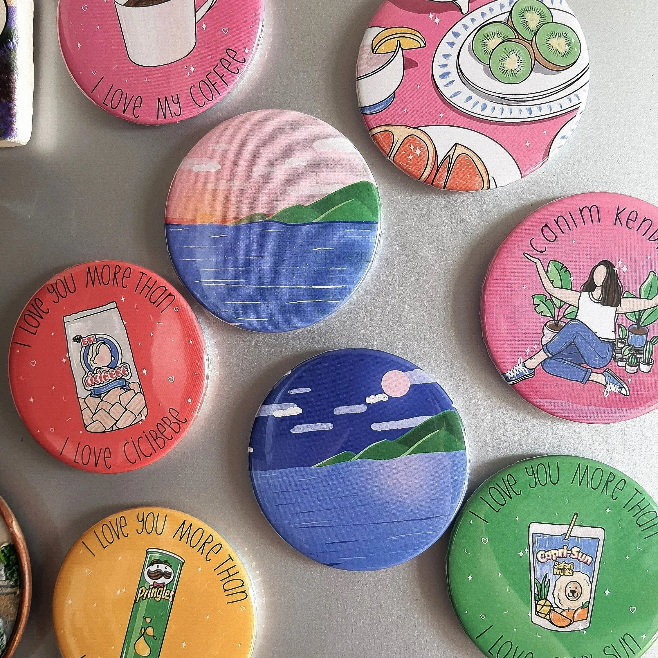 Colorful round pins with various designs and messages, including one that says 'I love my coffee' with a coffee cup illustration, another with a sunset over water, and others with motivational messages and illustrations of smoothies, kiwi, and a girl sitting among plants.