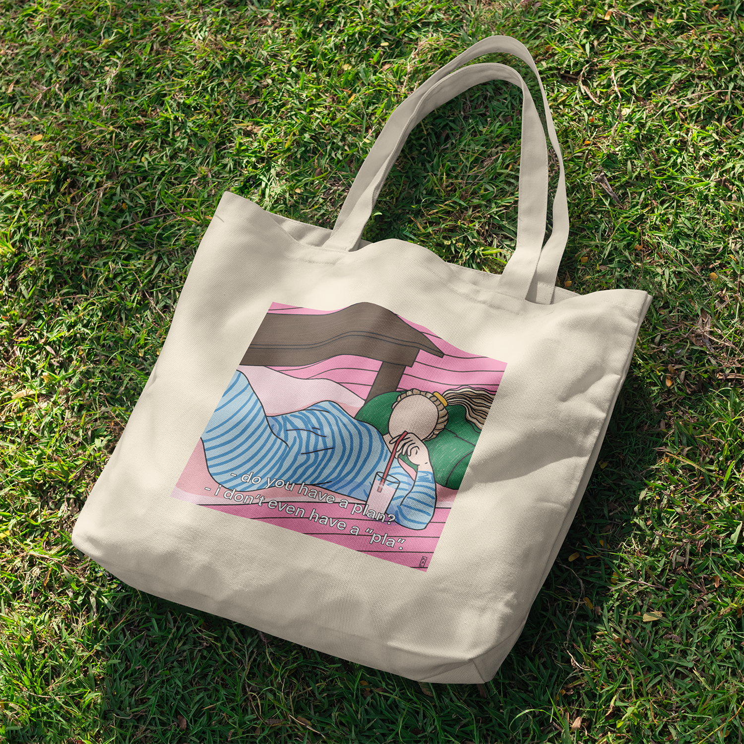 A beige tote bag lying on green grass with a colorful illustration of a person relaxing under a pink sky, drinking from a cup, with text that reads 'do you have a plan? - I didn't even have a 'pla'.