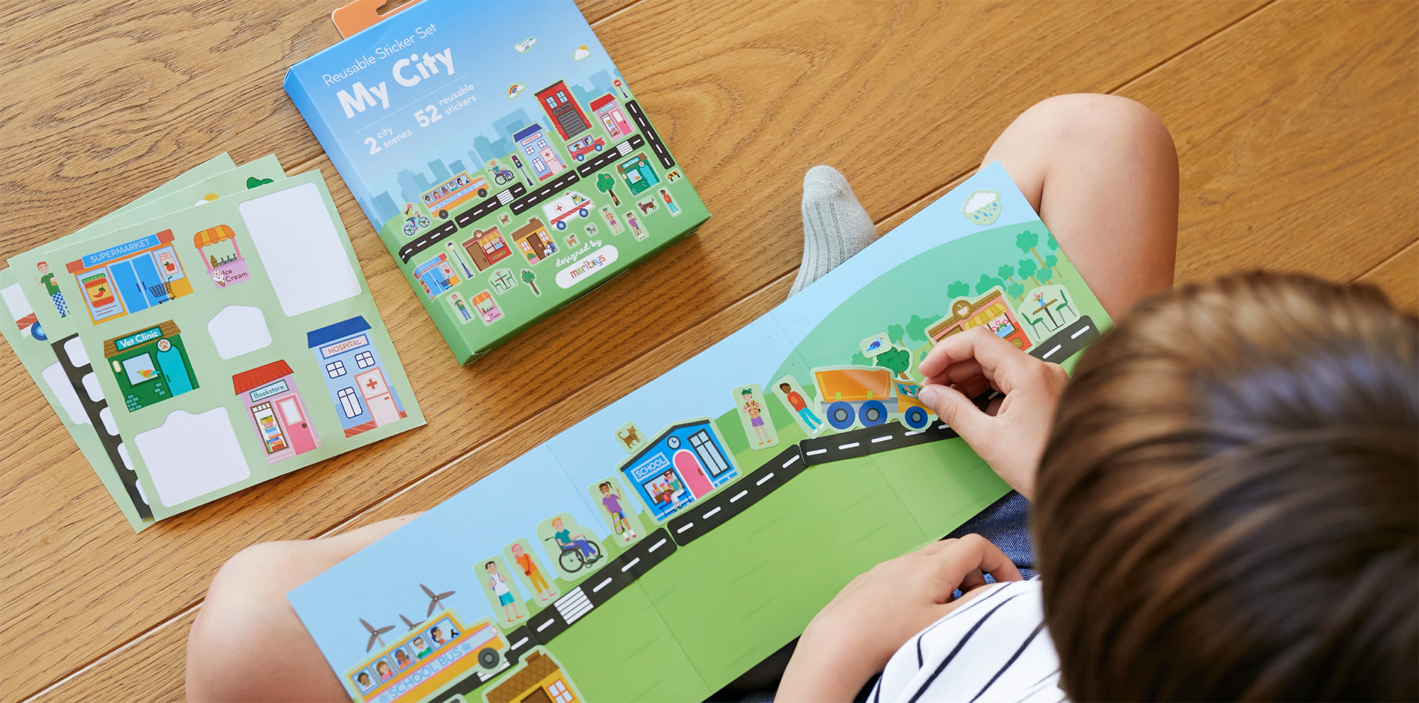 Children playing with a city-themed sticker set, placing stickers of buildings, vehicles, and people onto a large illustrated cityscape on a wooden table.