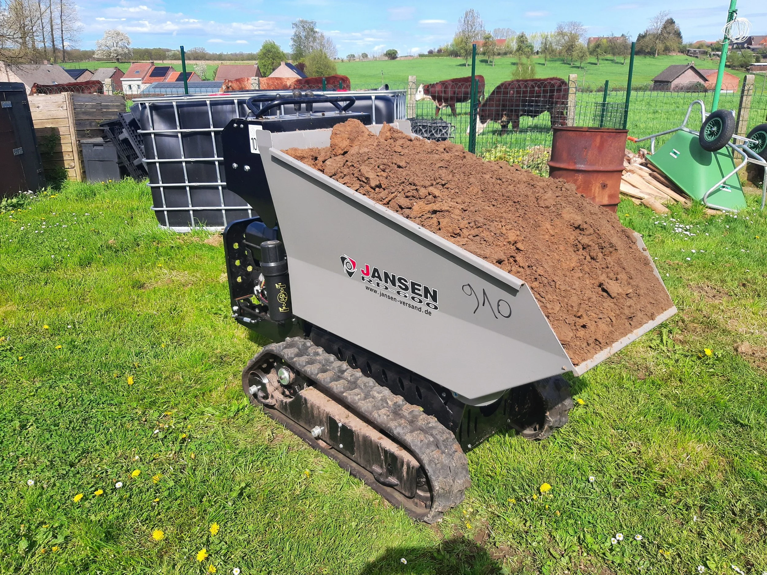 A small tracked construction vehicle filled with soil on a grassy yard with a fence, cows, and farm buildings in the background.