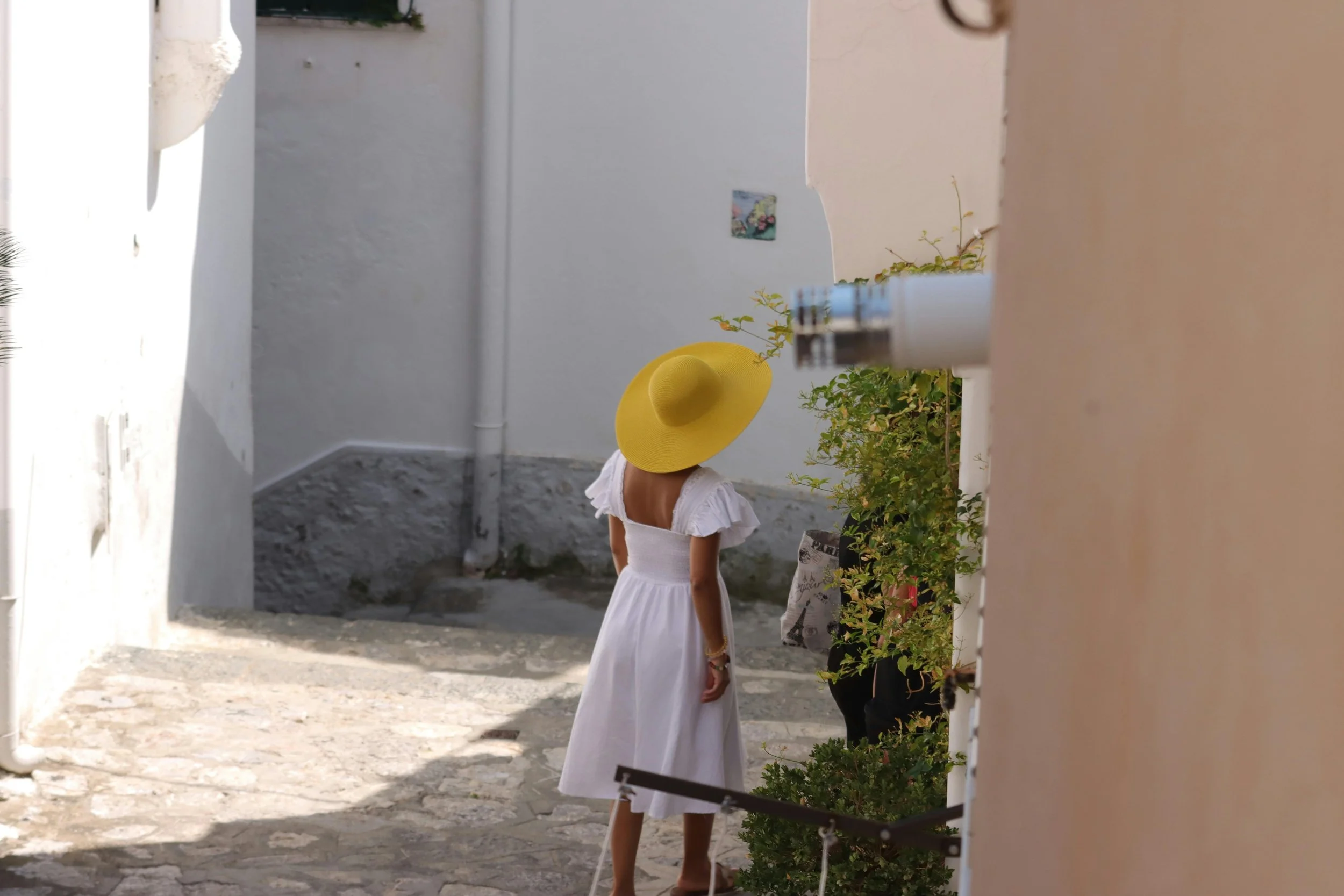 Woman in a white dress and large yellow hat standing in a narrow alleyway with white walls and stone pavement, facing away from the camera.