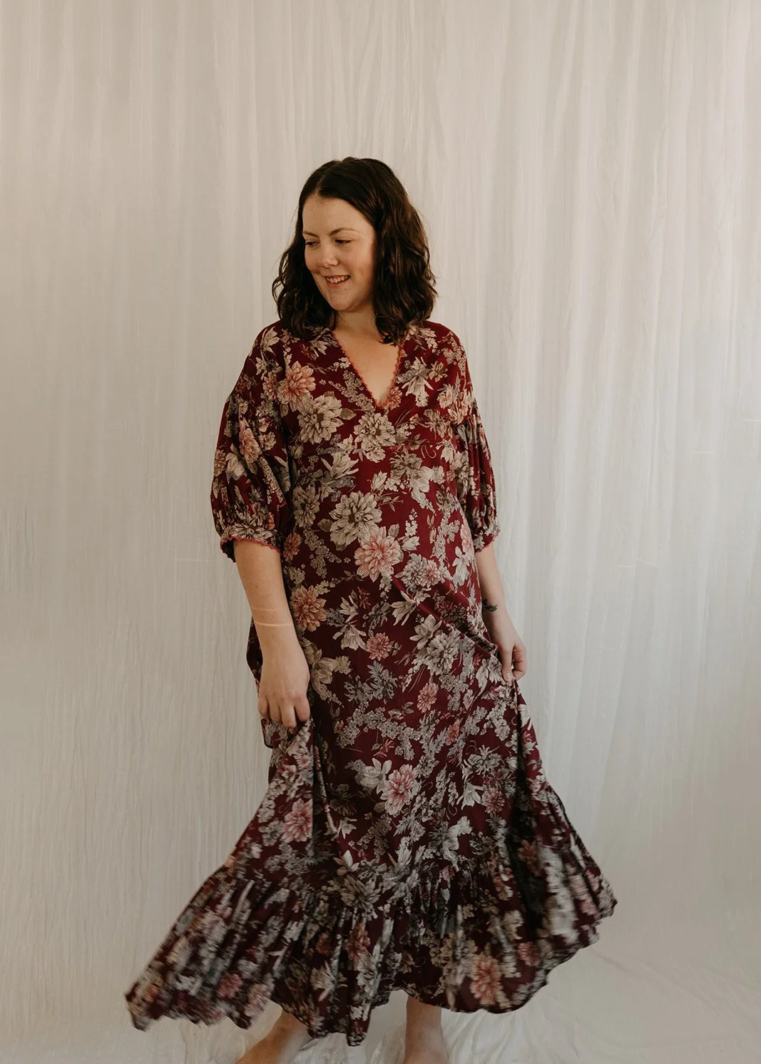 A woman wearing a long, floral dress with a maroon background and beige flowers, standing in front of a neutral-colored backdrop, looking down and smiling.