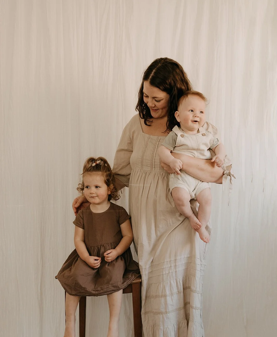 A woman holding a baby and standing next to a girl sitting on a chair, all posing in front of a plain light-colored background.
