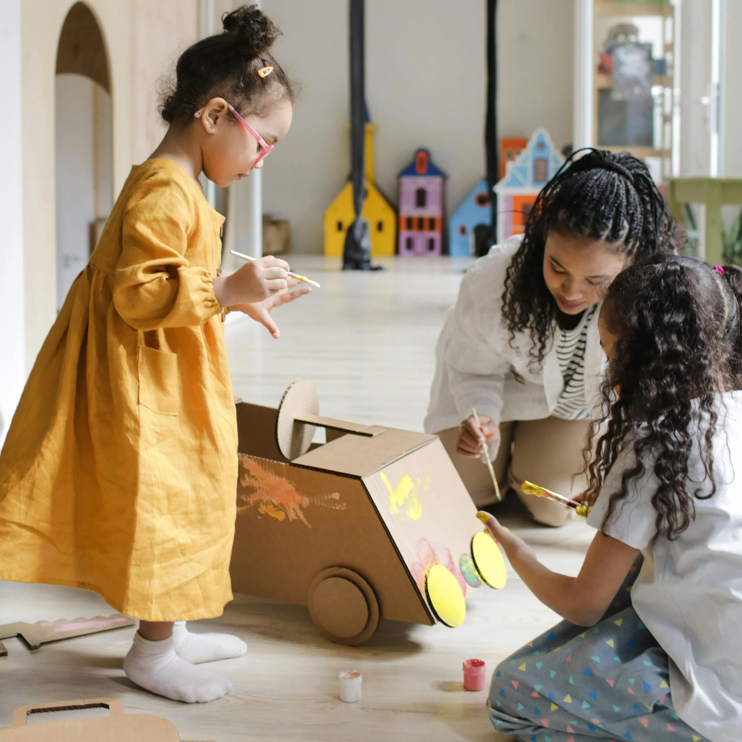 Three children and one adult painting a cardboard toy car in a classroom.
