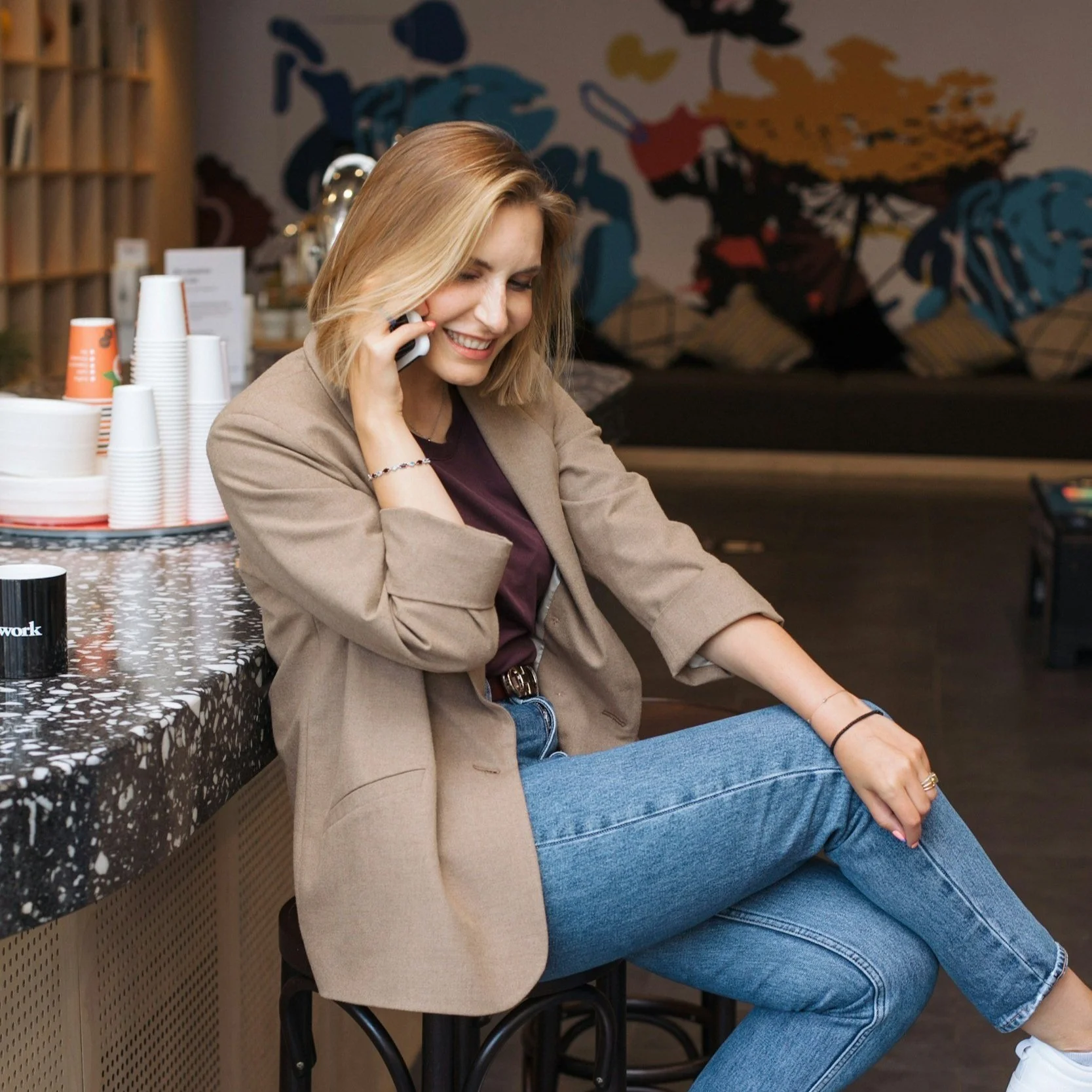 A woman with blonde hair smiling and talking on her phone while sitting at a bar or counter in a cafe or coffee shop. She is dressed in a beige blazer and blue jeans, and behind her are stacks of paper cups and a colorful wall art mural.