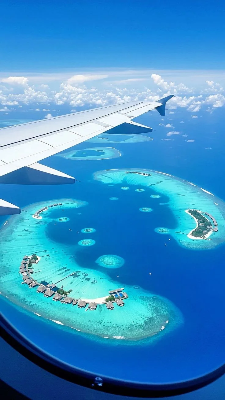 View from an airplane window showing a series of overwater bungalows and circular islands with turquoise water surrounding them, indicating a tropical vacation destination.