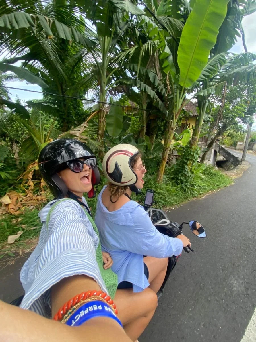 Two women riding a scooter on a paved road, surrounded by lush green tropical plants and trees, wearing helmets and smiling.