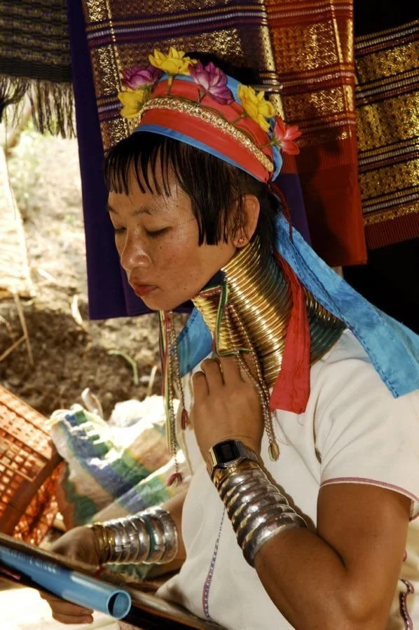 A person from the Kayan tribe, wearing traditional brass neck rings, colorful headpiece, and bracelets, reading a tablet.