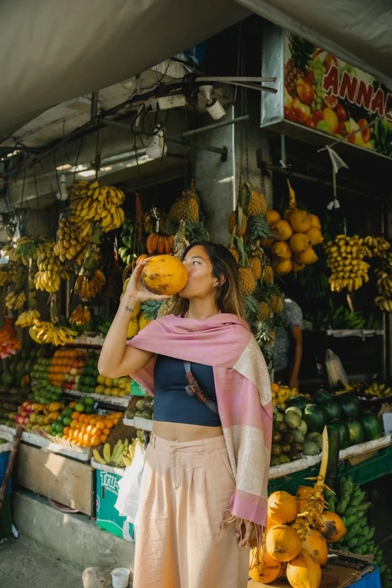 A woman drinking from a large papaya at a fruit stand filled with bananas, pineapples, and other fruits.