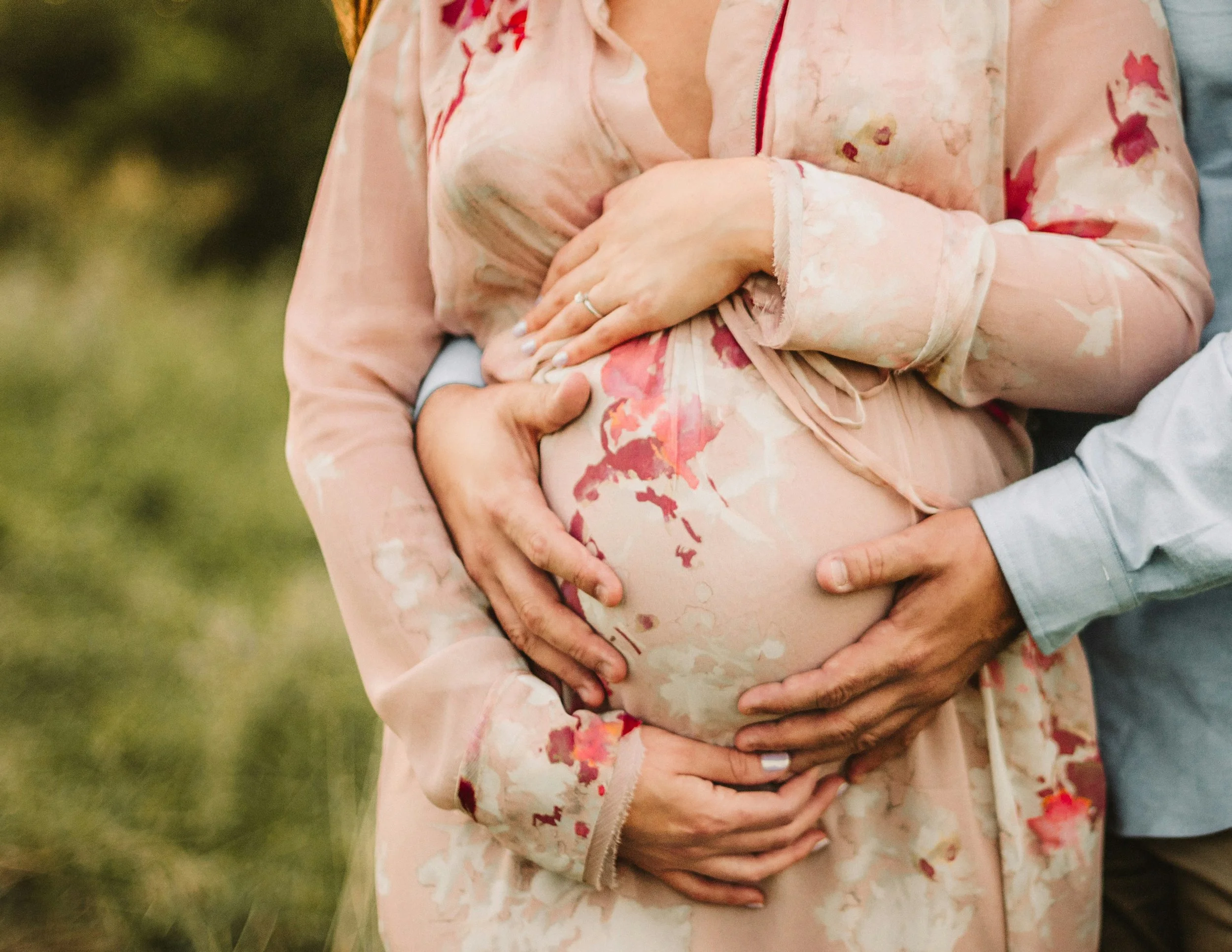 Close-up of a pregnant woman in a floral dress being affectionately touched on her belly by a partner's hands, outdoors.