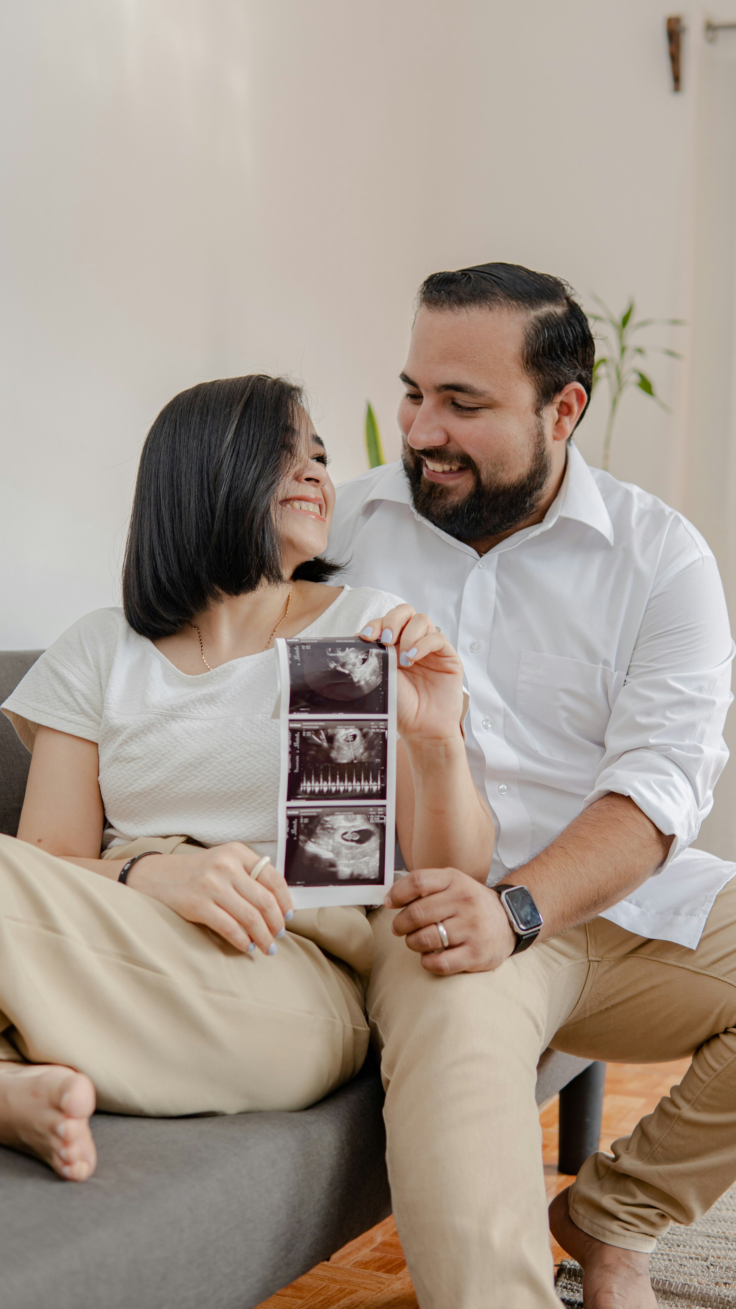 A smiling woman holding an ultrasound scan with a man sitting next to her, both appearing happy, inside a cozy room with plants in the background.