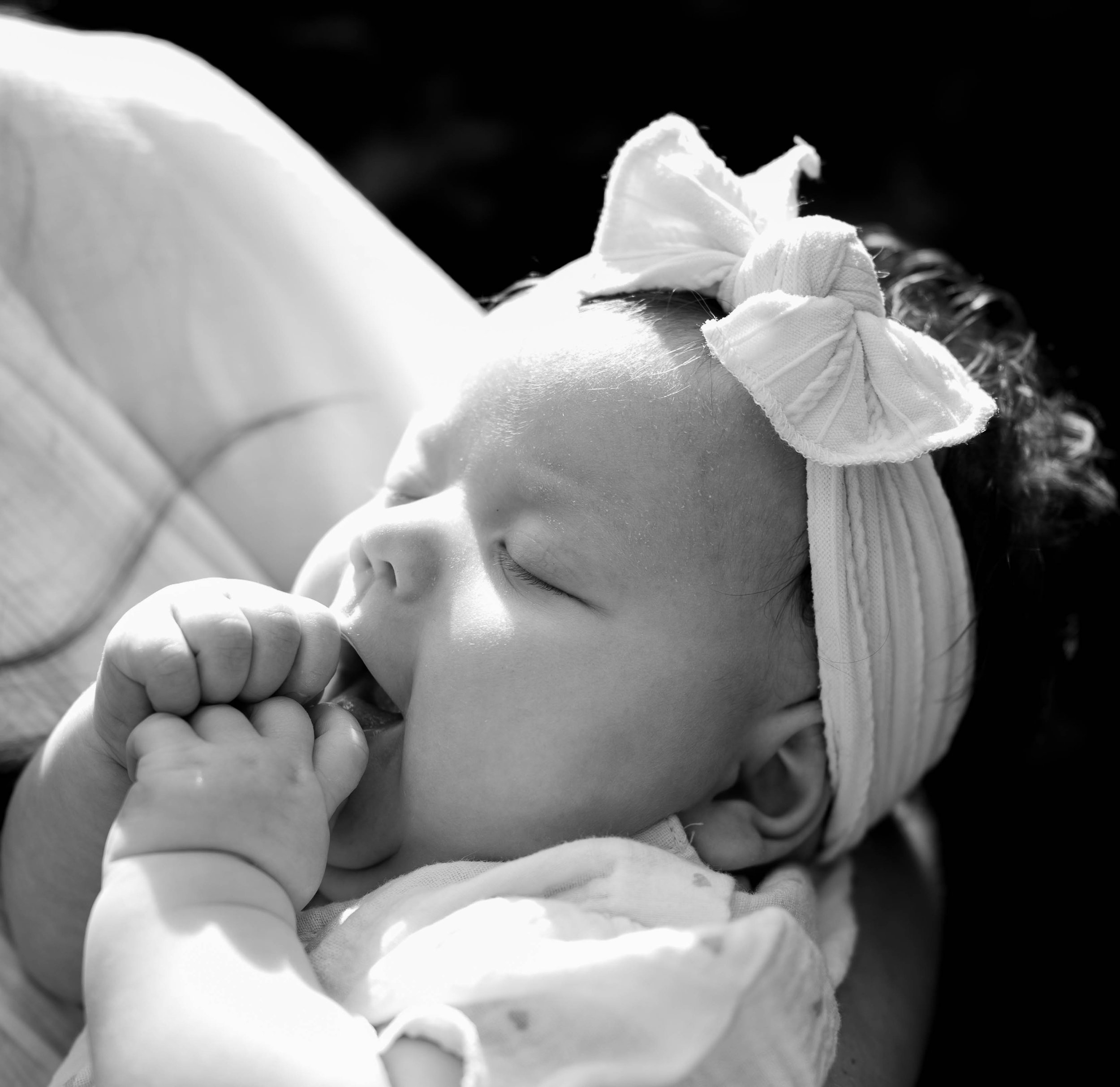 A sleeping baby girl with a large bow headband, resting in her mom's arms, with her hand near her mouth.