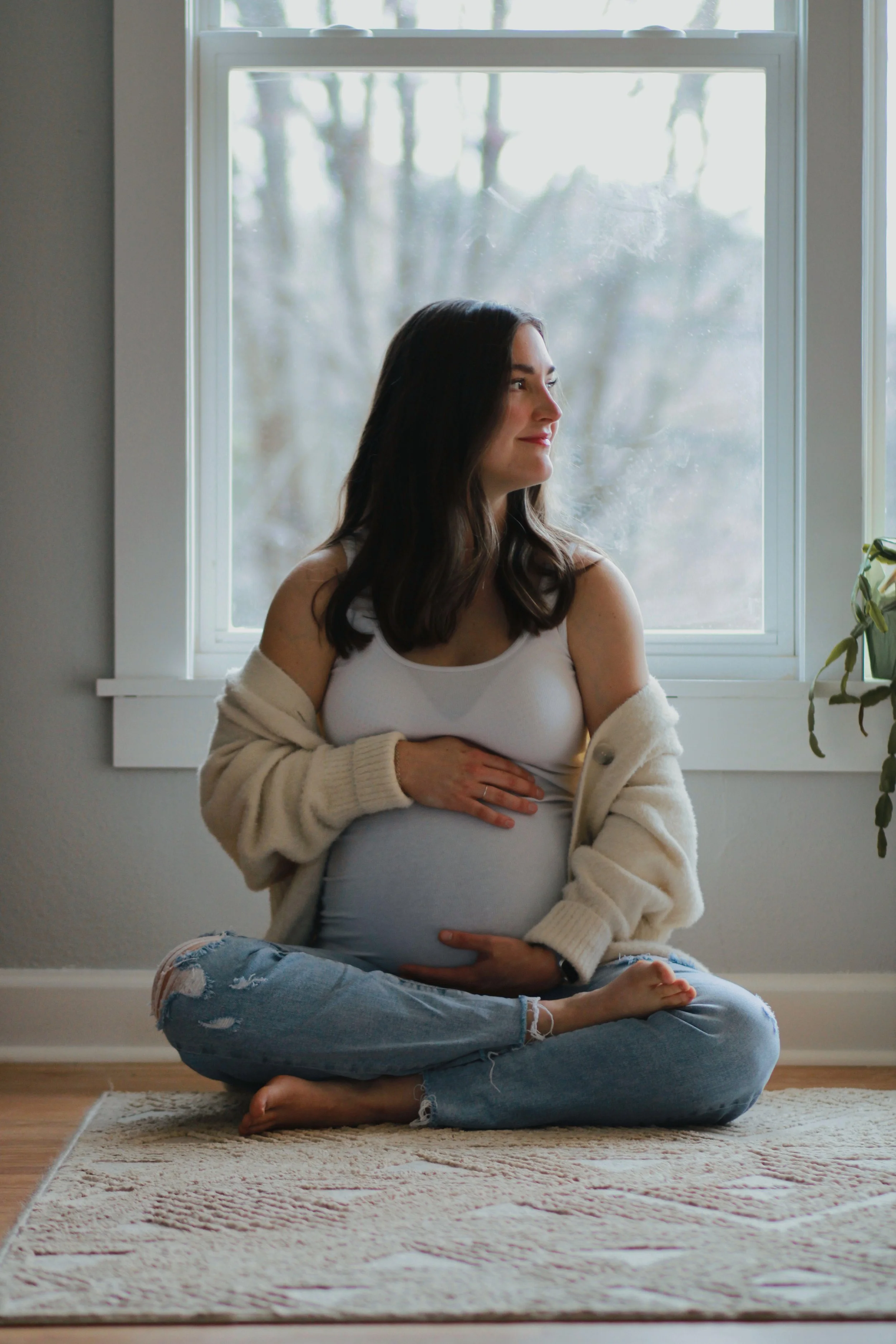 A pregnant woman sitting cross-legged on a rug in front of a window, cradling her belly, wearing a white tank top and ripped jeans, with a cozy cardigan draped over her shoulders, gazing out the window with a peaceful expression.
