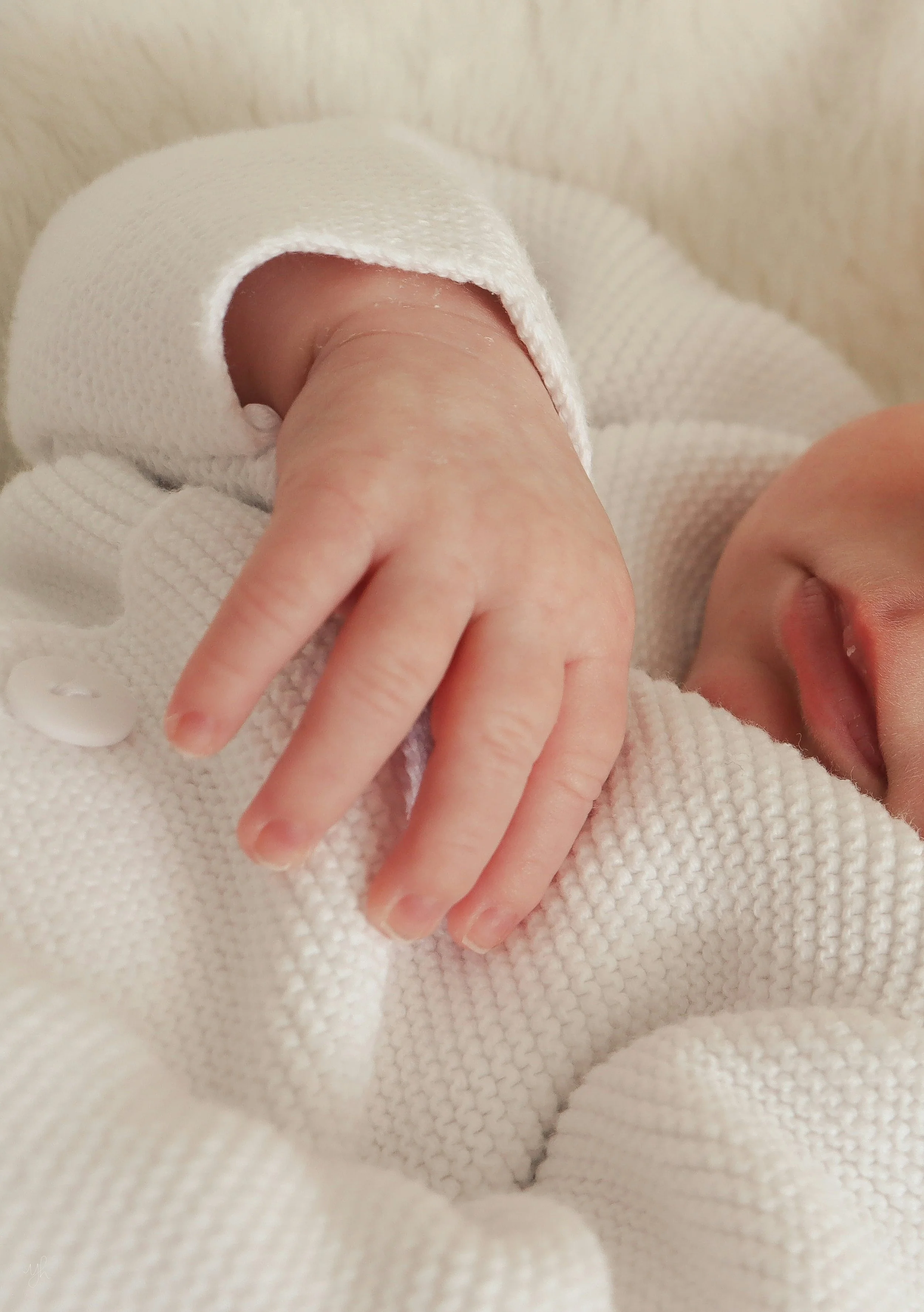 Close-up of a baby's hand resting on their chest, wearing a white knitted outfit, with the baby's face partially visible and lying on a soft surface.
