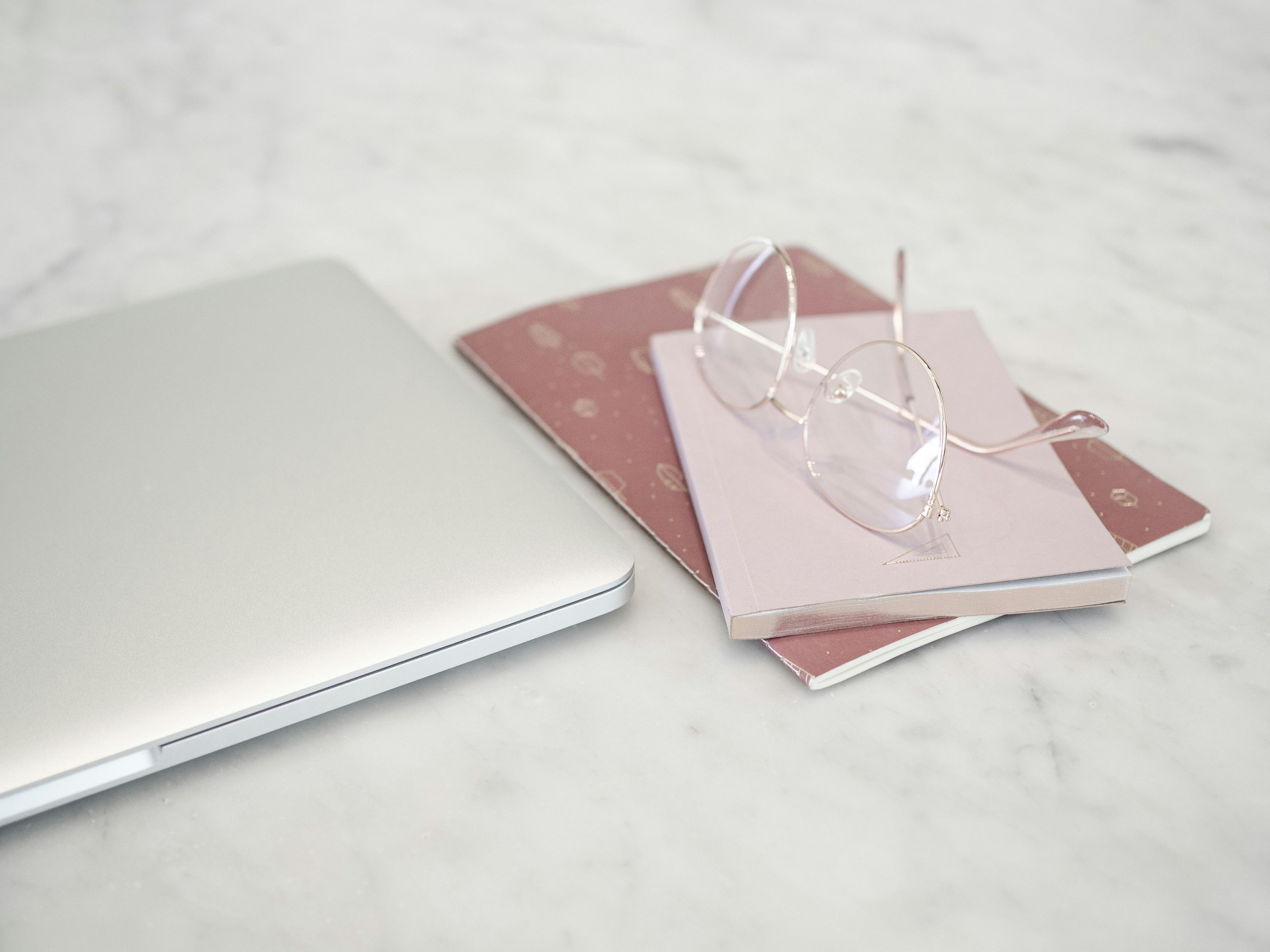 A closed silver laptop, two notebooks (one pink and one burgundy), and a pair of round, gold-rimmed eyeglasses placed on top of the notebooks on a white marble surface.