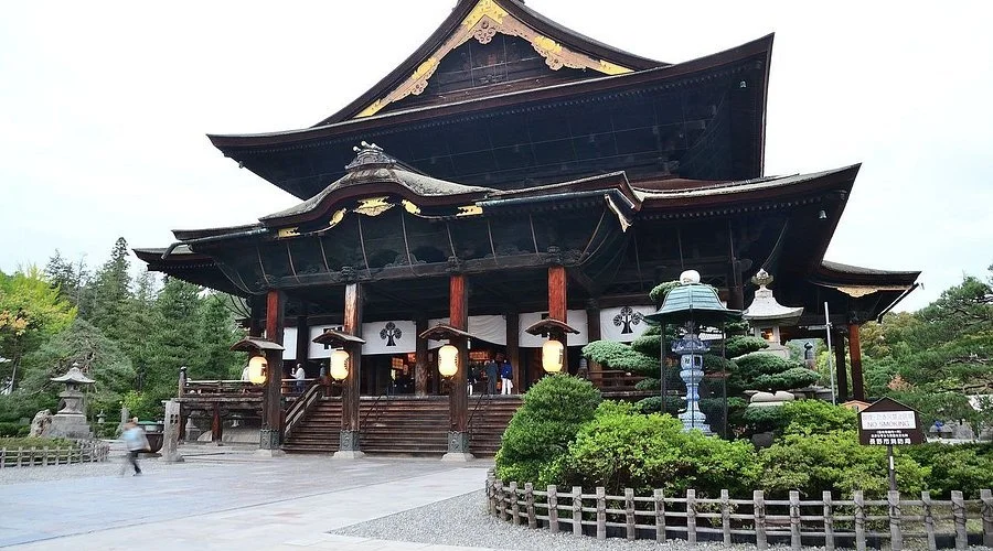 Traditional Japanese temple with curved wooden roof, lanterns, and well-maintained garden with bushes and trees.