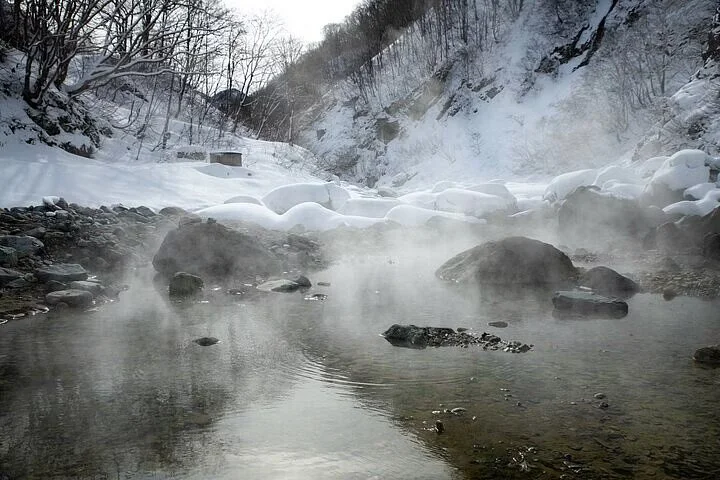 Steaming hot spring river with snow-covered banks and rocky shoreline, surrounded by leafless trees and rocky cliffs in a winter landscape.