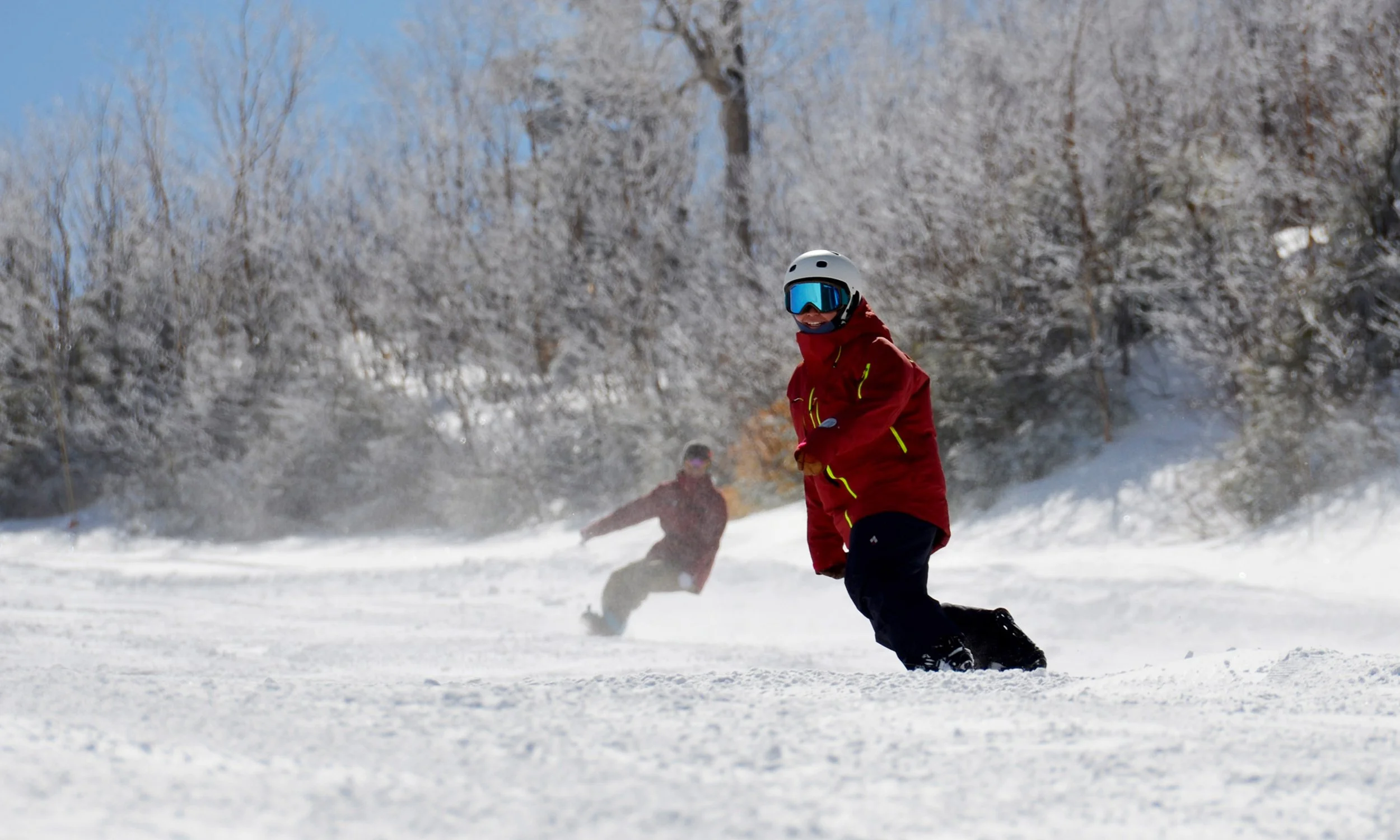 A person wearing a red jacket, black snow pants, and a white helmet with blue goggles is snowboarding on a snowy slope with frost-covered trees in the background. Another person in red can be seen in the distance.