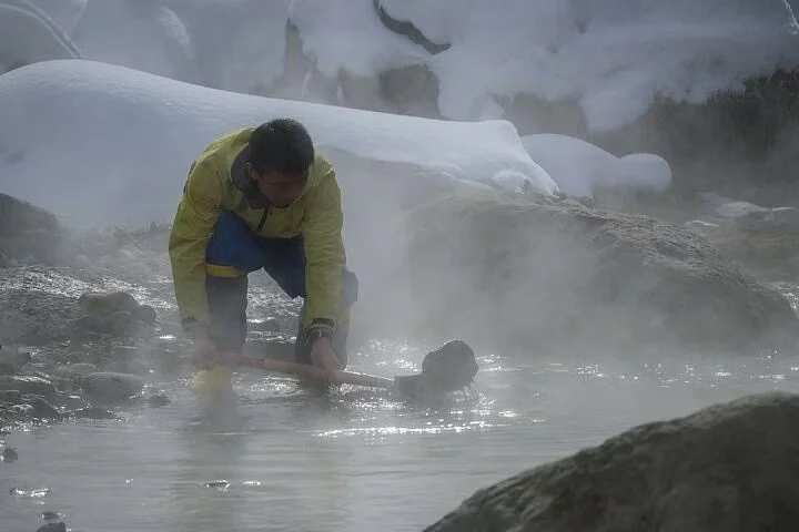 A person in a yellow jacket collecting or examining a black rock in a steaming hot spring or geyser area surrounded by rocks and steam.