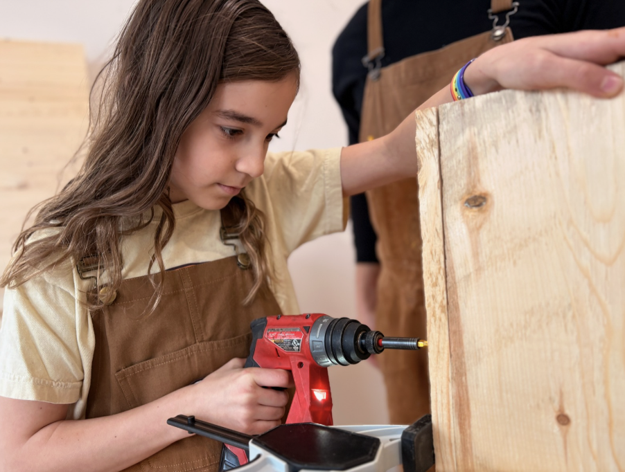 A child's small hand reaching into a wooden box of coins, with an adult's hand using pliers to pick coins from a compartment with multiple sections.