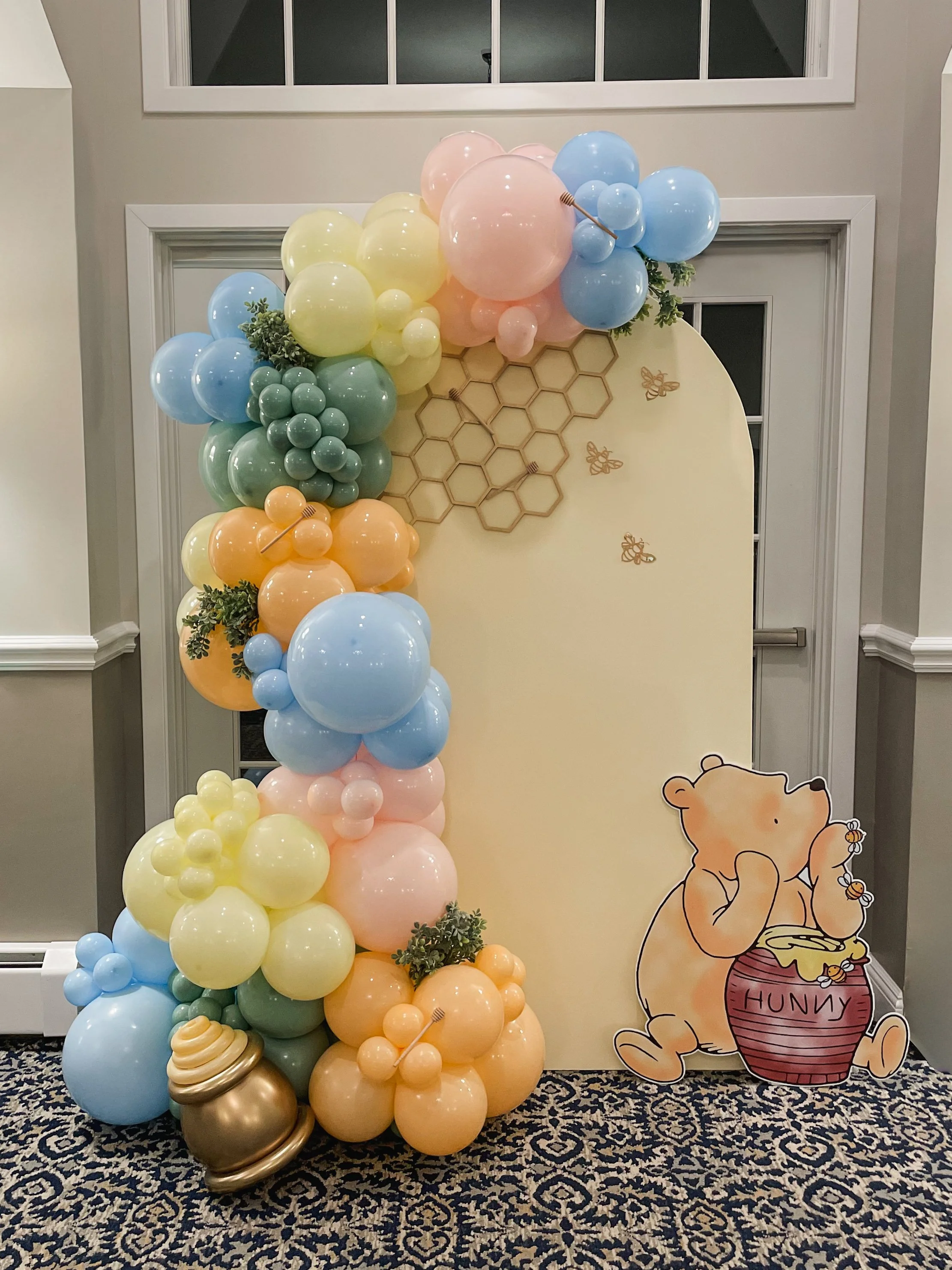 Decorative display featuring pastel-colored balloons in yellow, blue, green, pink, and peach, arrangement with artificial greenery, honeycomb and bee decor, and a Winnie the Pooh cutout with a honey jar labeled 'HUNNY'.